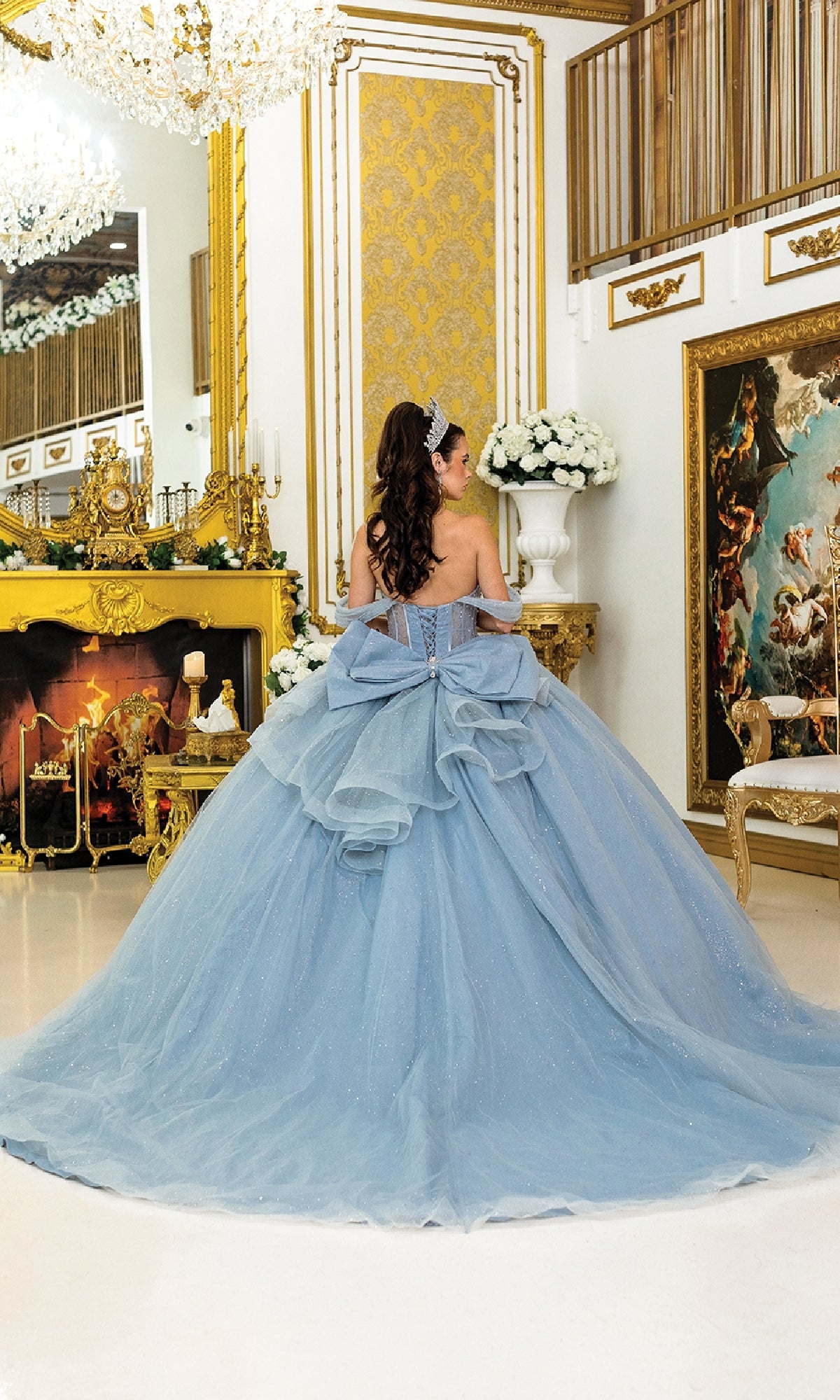 A woman in the Quinceanera Dress 1996 By Dancing Queen, featuring glitter tulle and a large bow, stands facing away in an ornate, gold-trimmed room with a fireplace, chandelier, and classical decor.