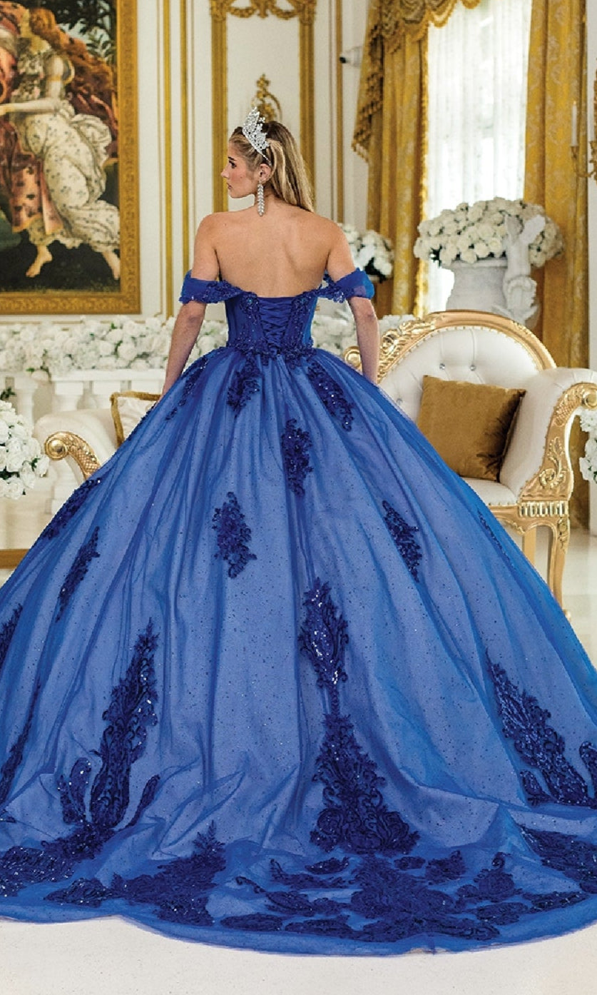 A woman in the Quinceanera Dress 1997 by Dancing Queen, featuring ornate blue sequin appliques and a tiara, stands with her back to the camera amid gold decor and white floral arrangements.