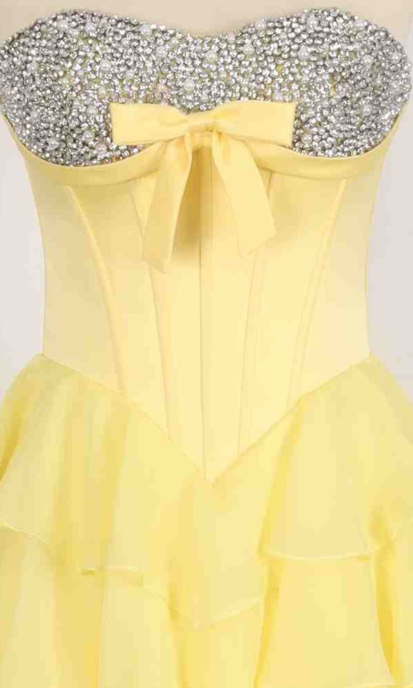 The PromGirl Exclusive 250203 is a strapless yellow ball gown with a layered skirt, fitted corset bodice adorned with silver rhinestones, and a charming yellow bow at the waist.