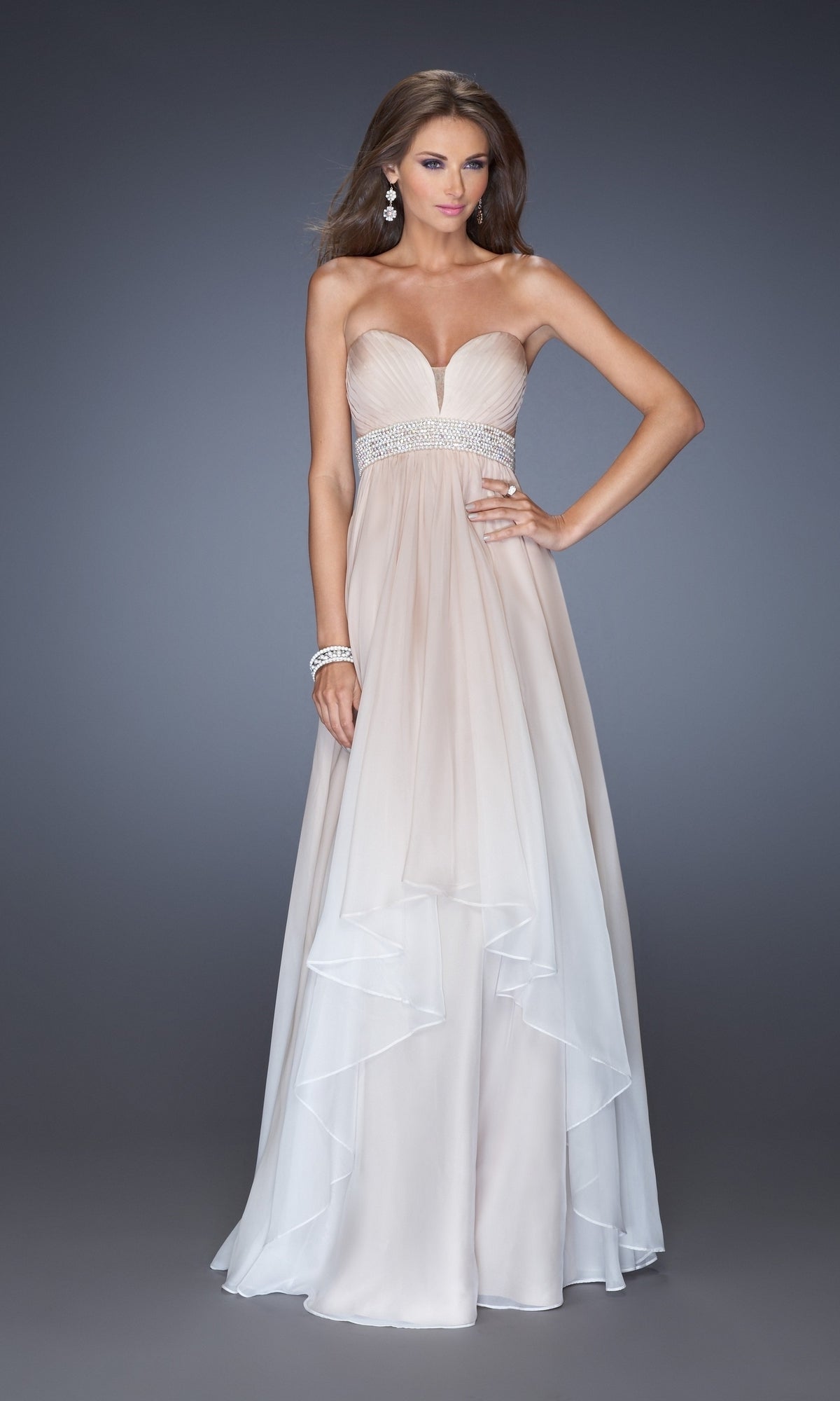 A woman models the La Femme Pleated Ombre Chiffon Prom Dress 20005, featuring a strapless design, jeweled waistband, and floor-length light beige skirt, as she poses gracefully against a plain gray background.