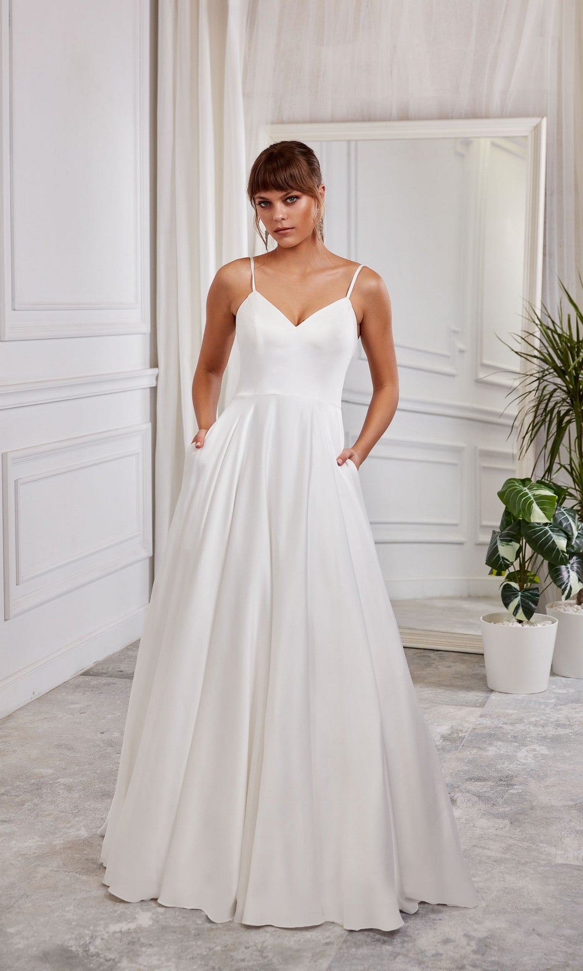 A woman wearing the Long White Dress: Aerie Couture 2000 in Diamond White, with thin straps and an a-line skirt, stands indoors beside a mirror and potted plants.