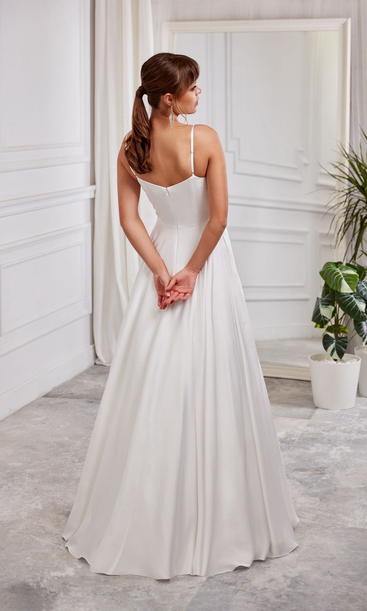 A woman wears the Long White Dress: Aerie Couture 2000, featuring an a-line skirt, standing with her back to the camera in a white room decorated with a large mirror, plants, and elegant paneling.
