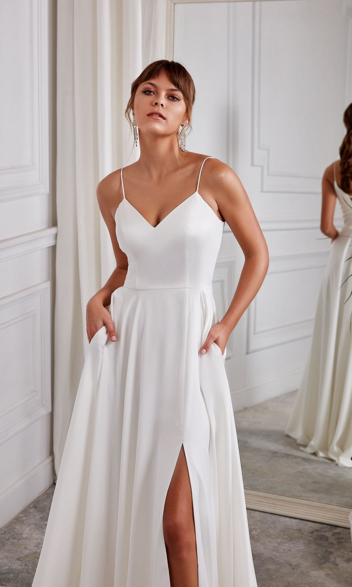 A woman wearing the Aerie Couture 2001 long white dress with thin straps, corset back, and thigh-high slit stands indoors by a mirror and white curtains.