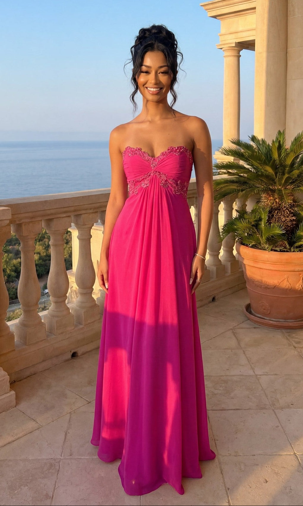 A woman in a hot fuchsia Strapless Long A-Line Prom Dress (La Femme 20042) stands on a stone balcony, with the ocean and lush greenery in the background.