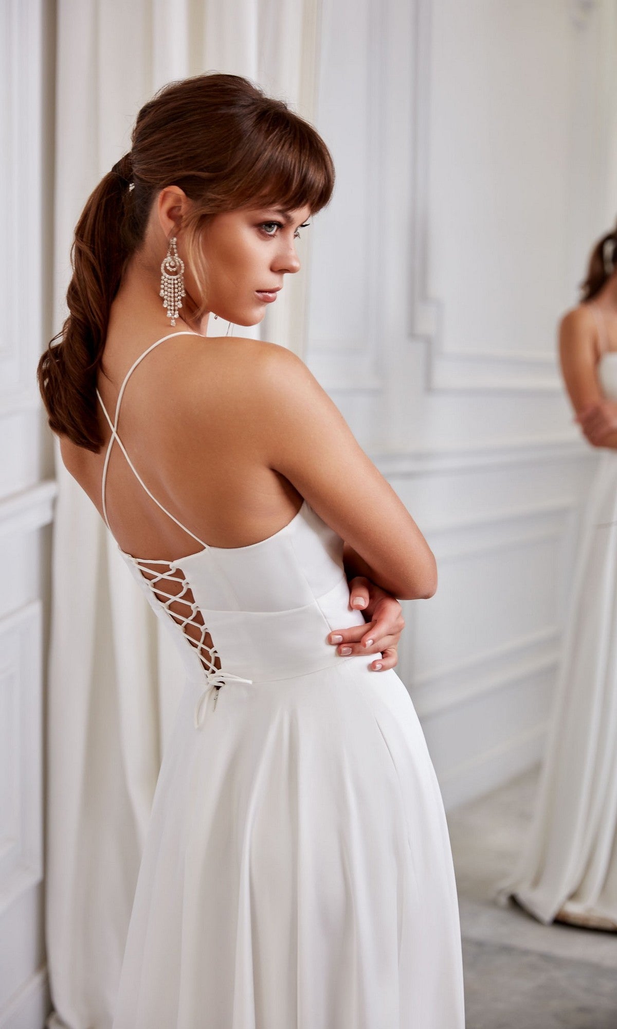 A woman wearing the Long White Dress: Aerie Couture 2005, an A-line gown with crisscross straps and a corset back, stands in a white room and looks over her shoulder.
