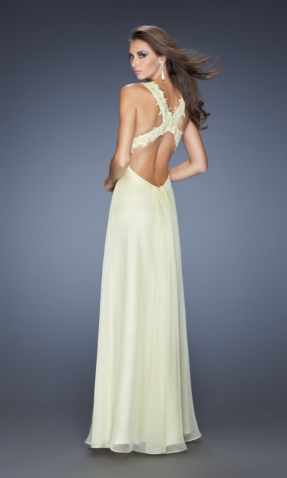 Model wears the La Femme Empire-Waist Ruched Chiffon Prom Dress 20134 in pale yellow, featuring an open back and lace shoulder straps, standing against a plain gray background.