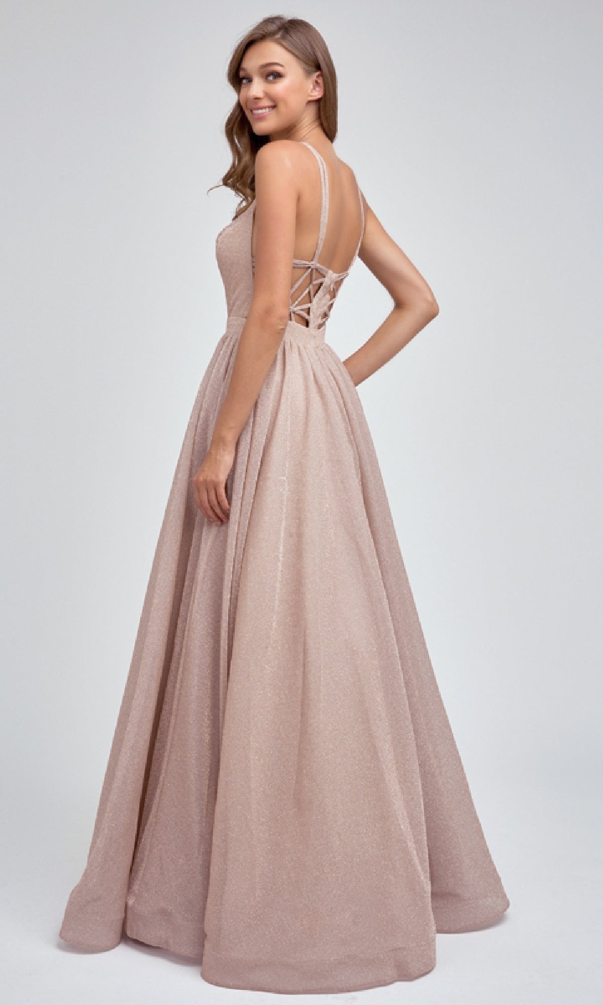 A woman poses and smiles over her shoulder against a plain background, wearing the Juliet 201 long prom dress—a sleeveless, light pink, floor-length gown with an A-line skirt and lace-up back detail.