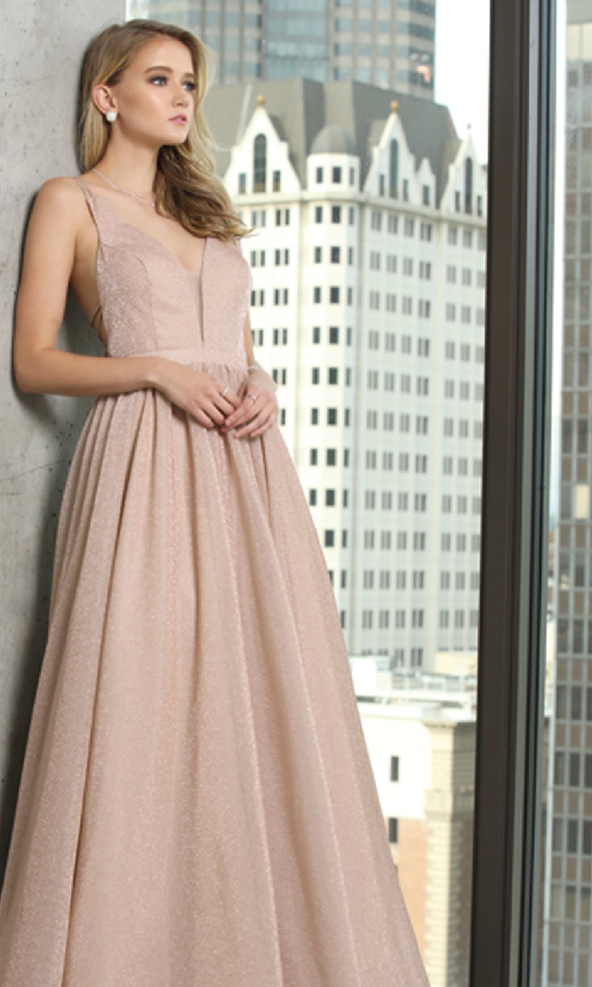 Wearing the Juliet 201 Long Prom Dress, a woman in a sleeveless blush gown with an elegant cut-out back stands by a window, city skyline and tall buildings visible behind her.