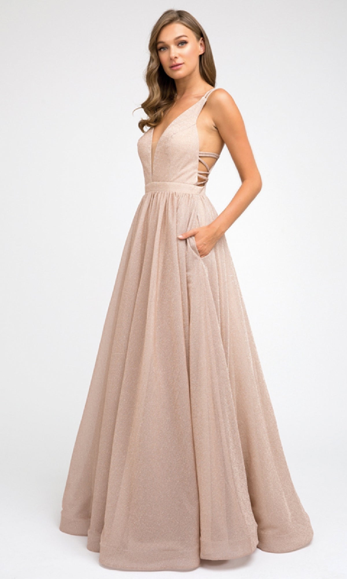 A woman poses against a white background in the Juliet 201 Long Prom Dress, featuring a sleeveless, floor-length blush pink design with a deep V-neckline, side cutouts, and an elegant A-line skirt.