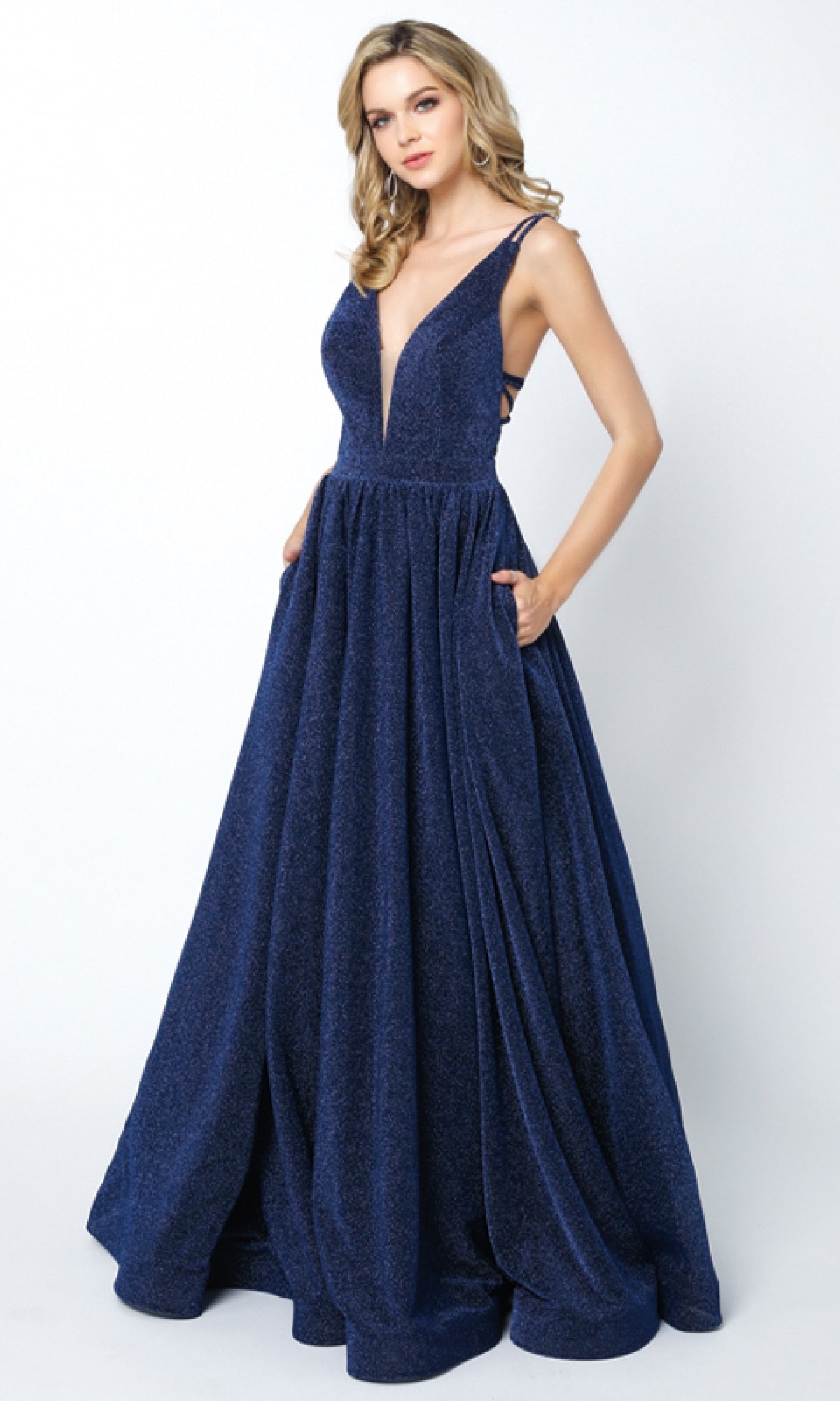 A woman wearing the Long Prom Dress: Juliet 201—a deep blue, sleeveless, floor-length gown with a deep V-neckline, a-line skirt, pockets, and subtle cut-out back—stands against a plain white background.