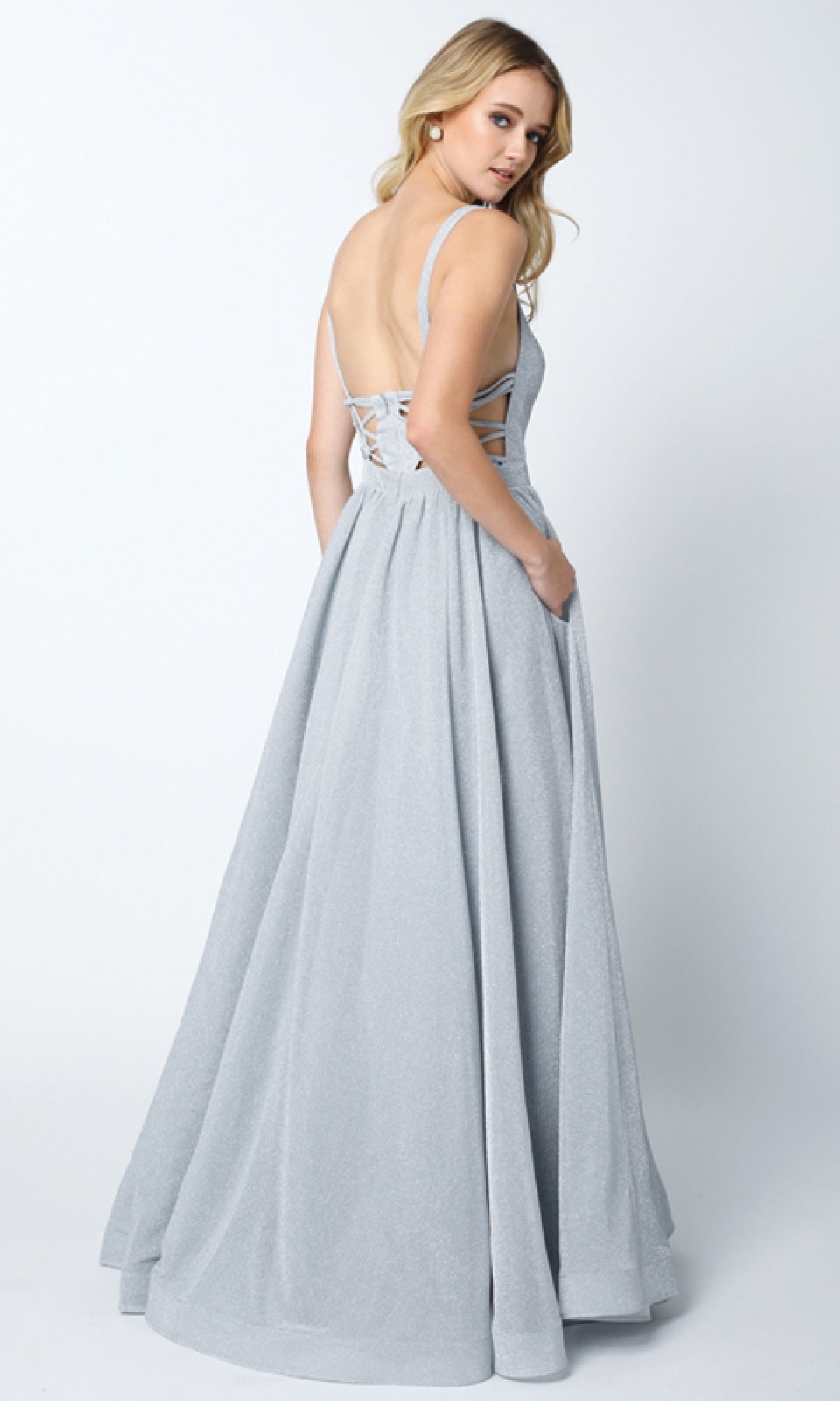 A woman in the Juliet 201 Long Prom Dress, a light gray floor-length gown with an a-line skirt, lace-up detailing, and a cut-out back, stands and looks over her shoulder.