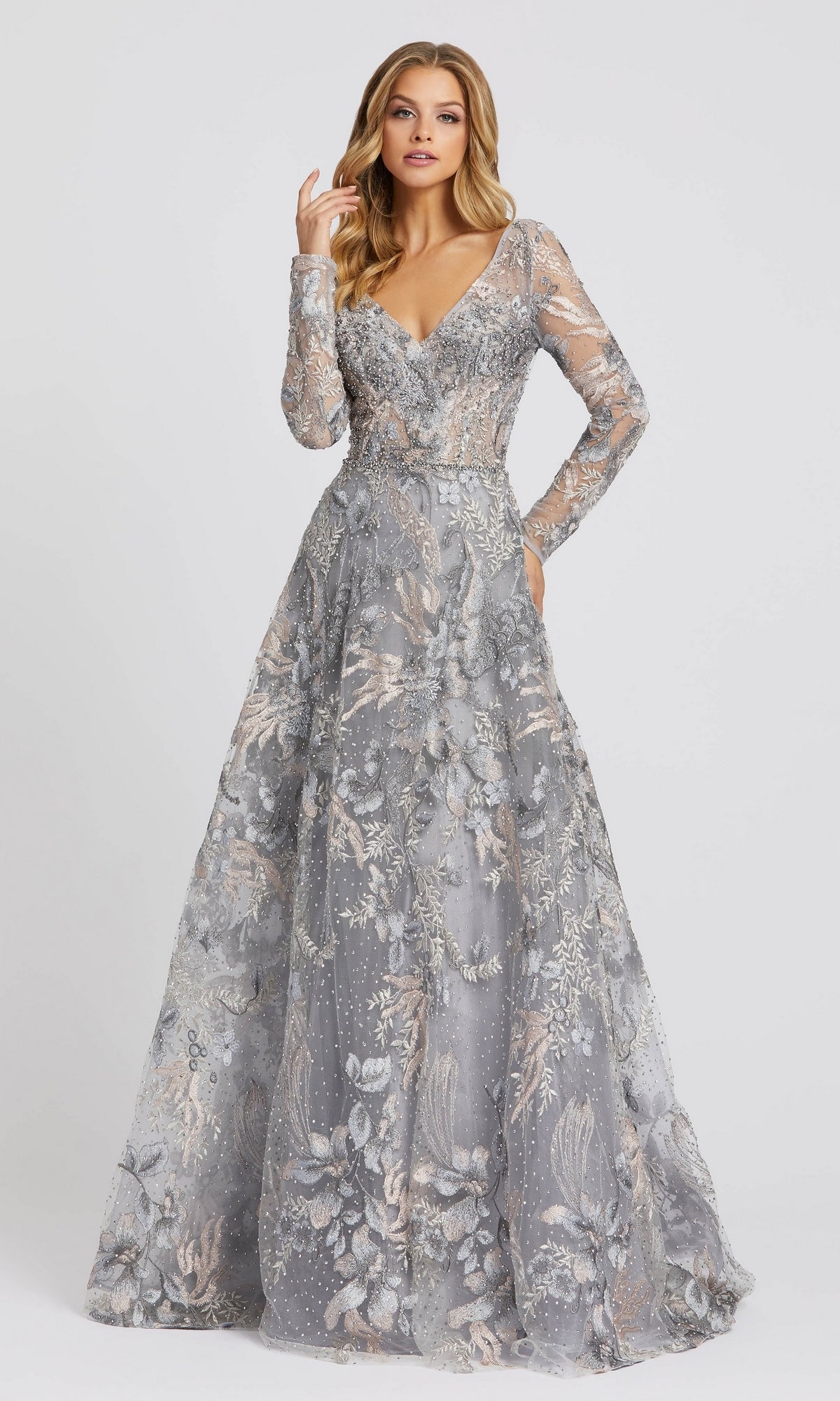 A woman wears the Mac Duggal 20214, a floor-length silver-gray formal dress with long sleeves, intricate floral embroidery, and a deep V-neckline, standing against a plain light background.
