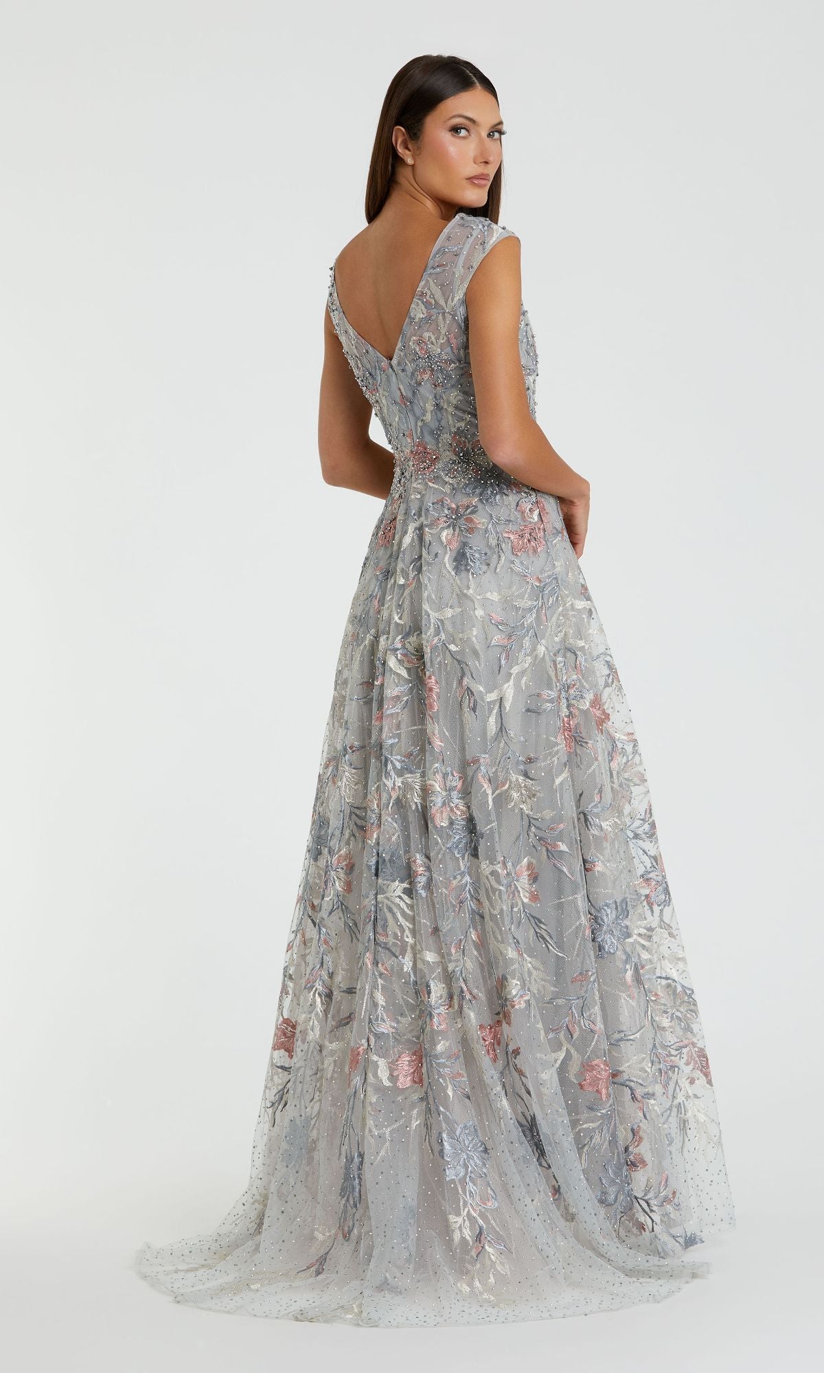 A woman models the Mac Duggal 20263 Cap-Sleeve Long Print Prom Dress, featuring floral embroidery, with her back partially turned to the camera against a plain backdrop.