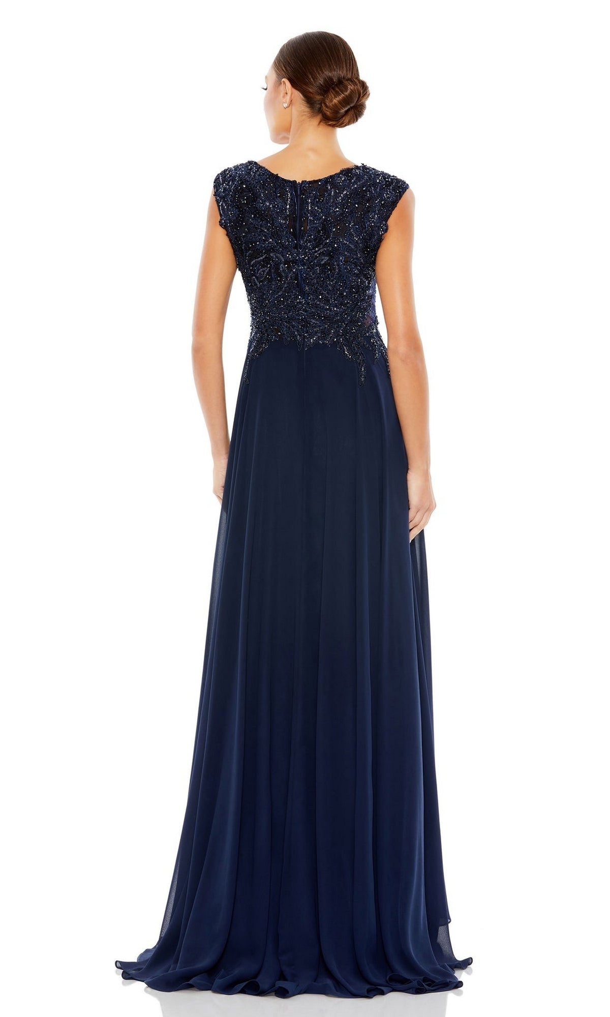 A woman with hair in a low bun faces away, wearing the Mac Duggal 20264 Beaded-Bodice Long Formal Dress in navy blue, featuring cap sleeves, a beaded v-neck bodice, and a flowing chiffon skirt.