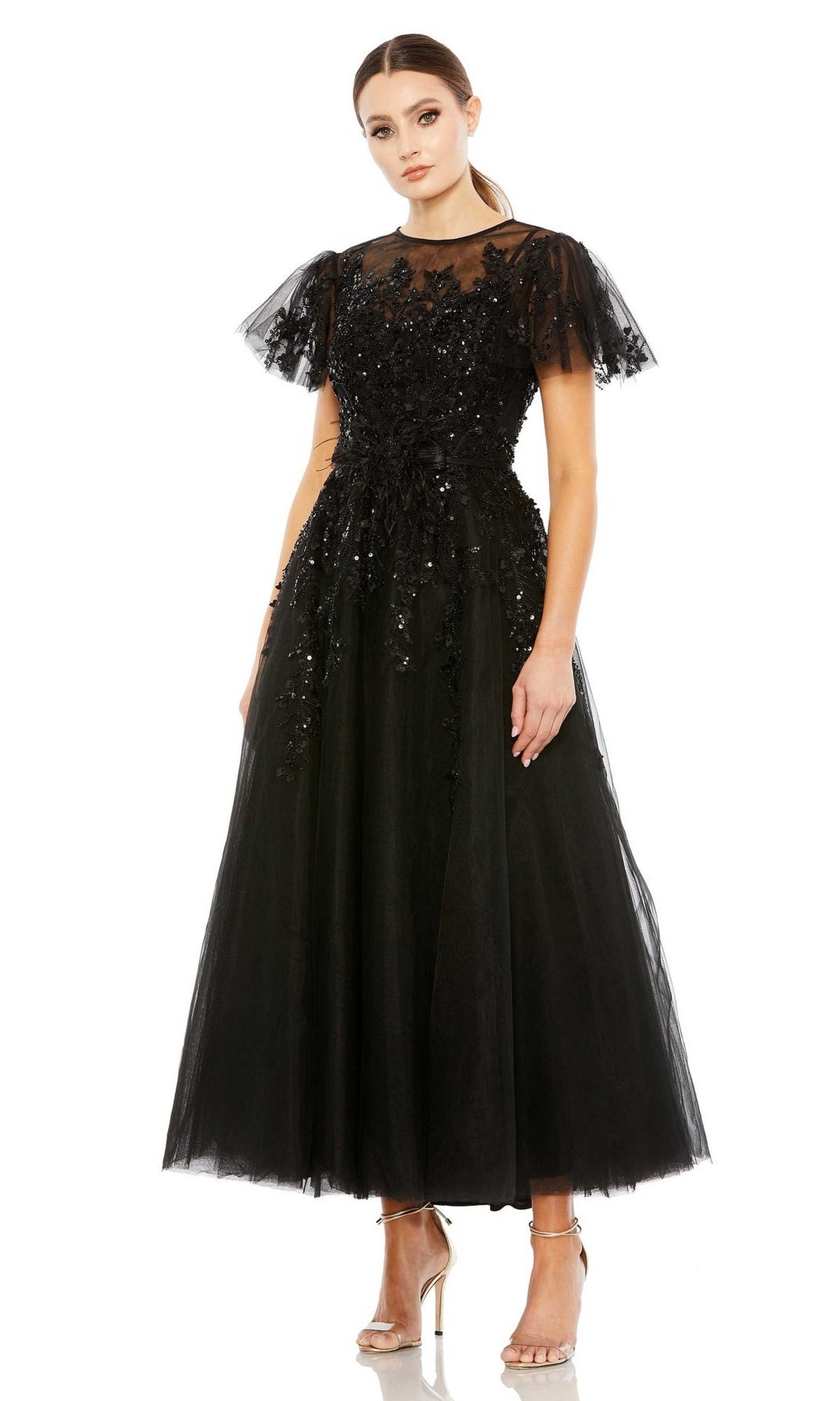 A woman stands against a plain white background wearing the Long Formal Dress 20362 by Mac Duggal, a black tea-length tulle gown with flutter sleeves, floral appliqué details, and metallic high-heeled sandals.