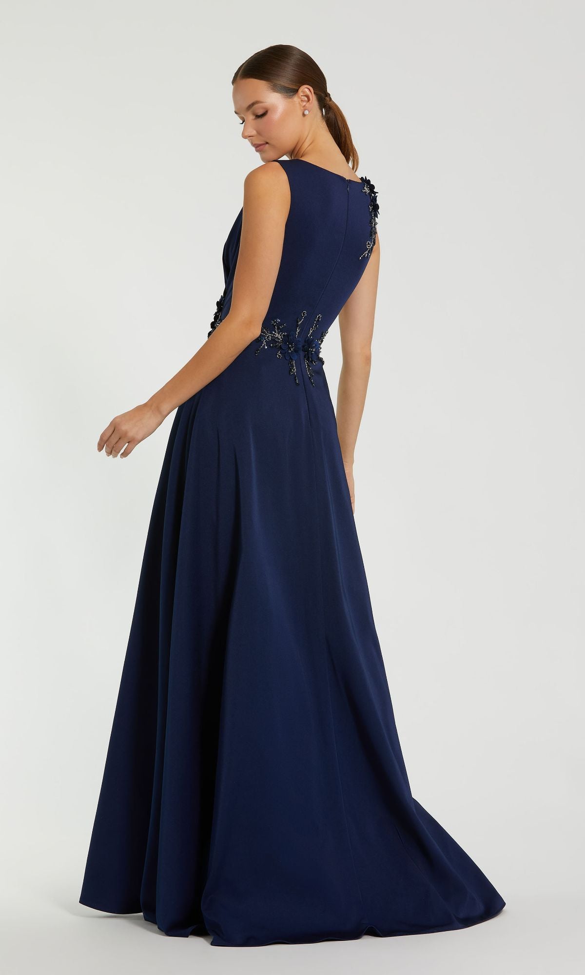 A woman models the Mac Duggal 20365 Sleeveless Long Formal Dress, a floor-length navy blue gown with floral back embellishments, as she stands sideways against a plain light background.