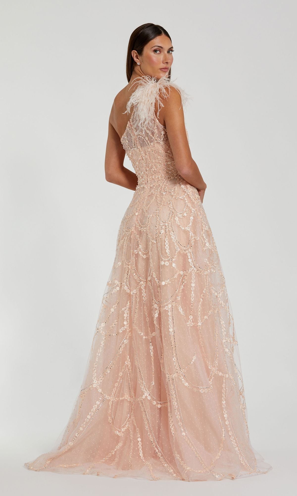 A woman faces away in the Mac Duggal 20389 Beaded One-Shoulder Prom Gown—a light pink, floor-length dress with sequin patterns and feathered shoulder details—posed against a plain white background.