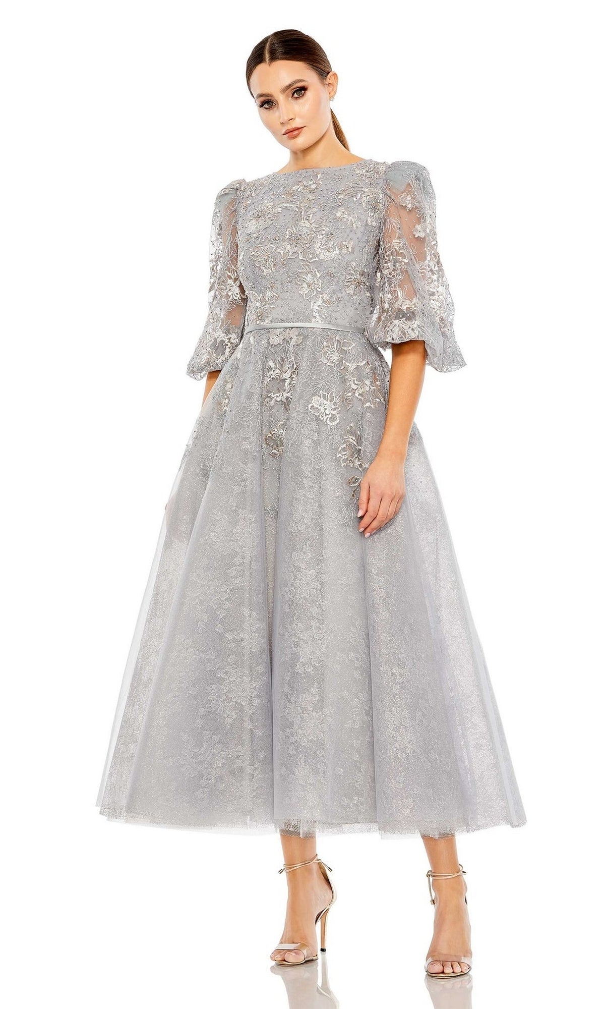 A woman models the Long Formal Dress 20418 by Mac Duggal, a light gray tea-length dress with puff sleeves and floral embroidery, paired with silver high-heeled sandals against a white backdrop.