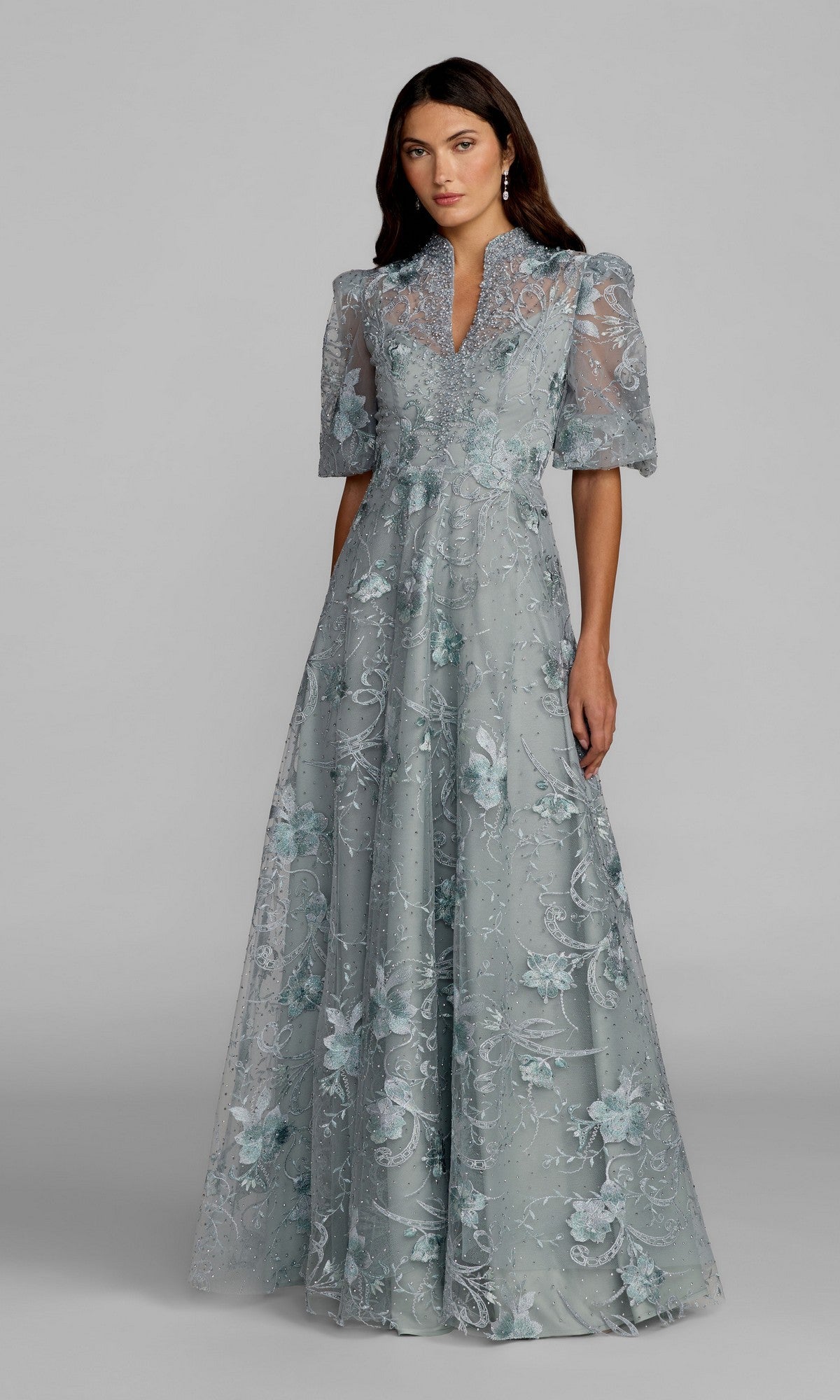 A woman stands against a plain background, wearing the Mac Duggal 20452 Short Sleeve Beaded Formal Gown in French Blue—a floor-length dress featuring floral embroidery and sheer short sleeves for an elegant look.