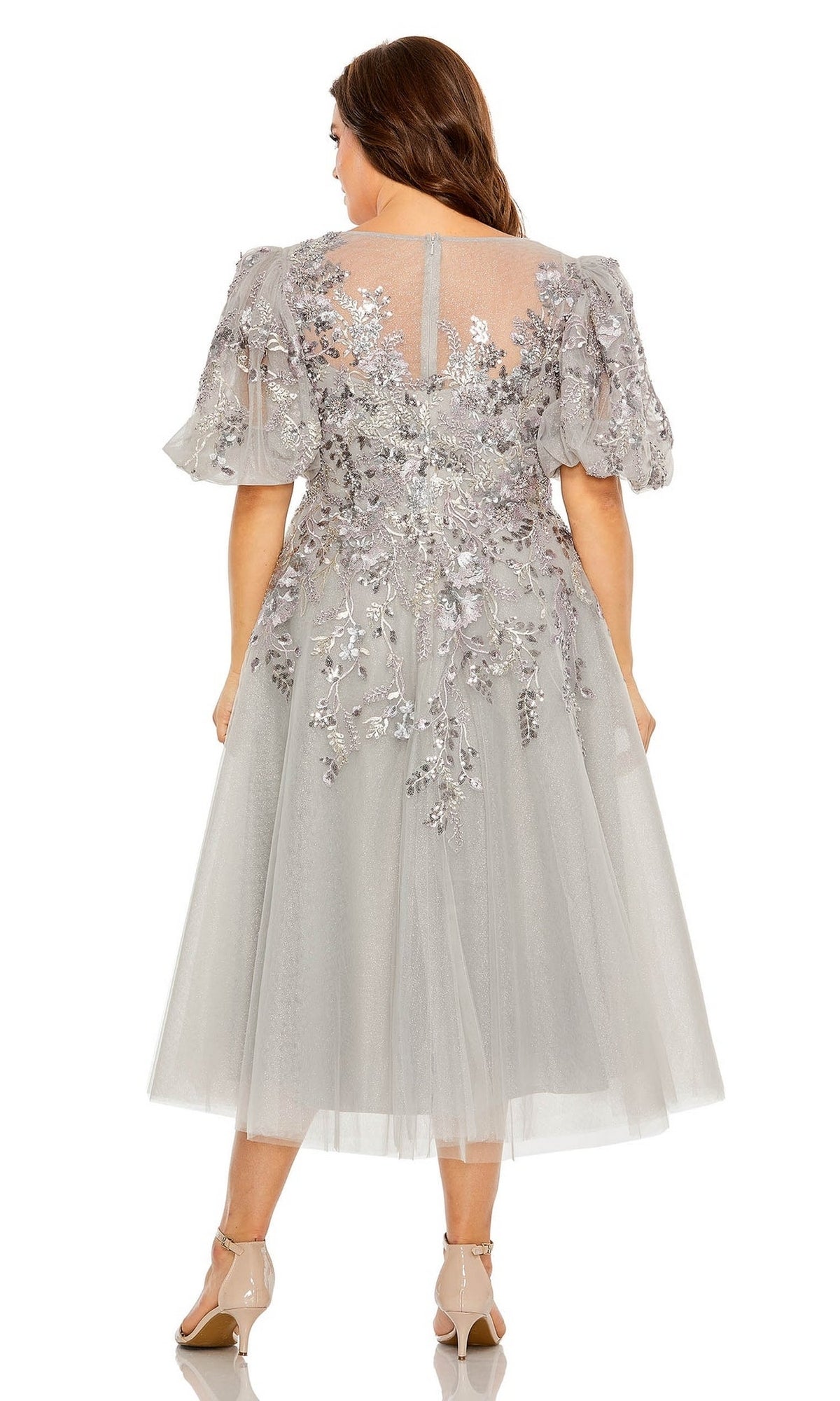 A woman faces away in the Plus-Size Semi-Formal Dress 20475 by Mac Duggal—a light grey, floral embroidered midi A-line with short puff sleeves and a tulle skirt—paired with beige heels and long brown hair flowing down.