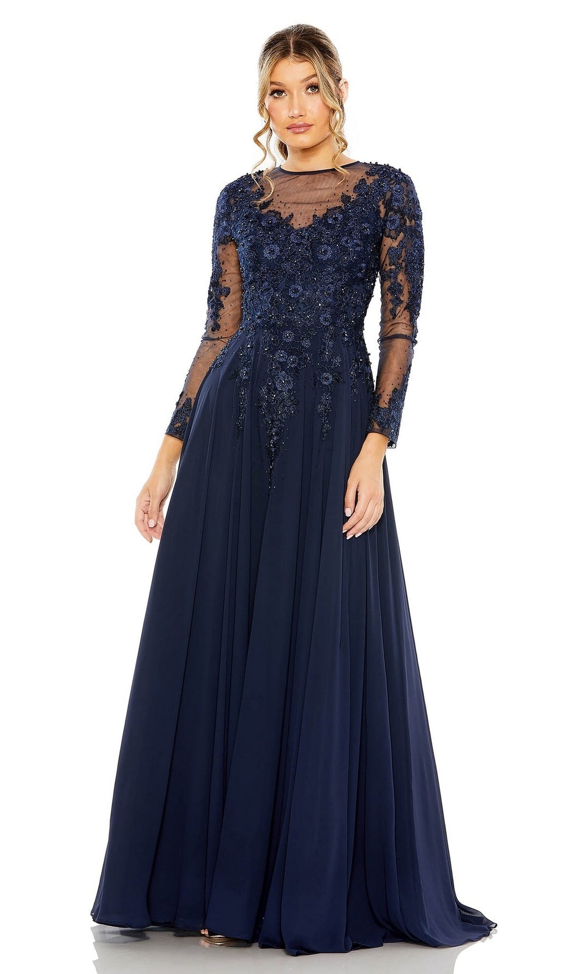A woman wearing the Mac Duggal 20478 long sleeve formal gown, featuring a navy blue A-line silhouette with sheer sleeves, floral embroidered bodice, and flowing skirt—a stunning mother of the bride dress.