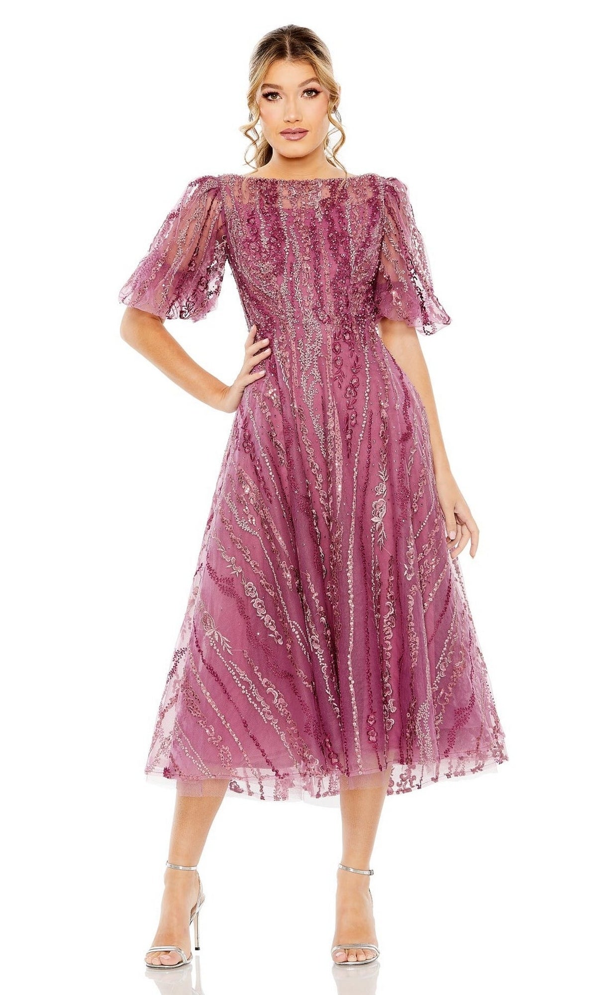 A woman wears the Mac Duggal 20485 Embroidered Wedding-Guest Dress, a mauve midi with sheer short puff sleeves, detailed embroidery, and sequin accents, styled with silver high-heeled sandals.