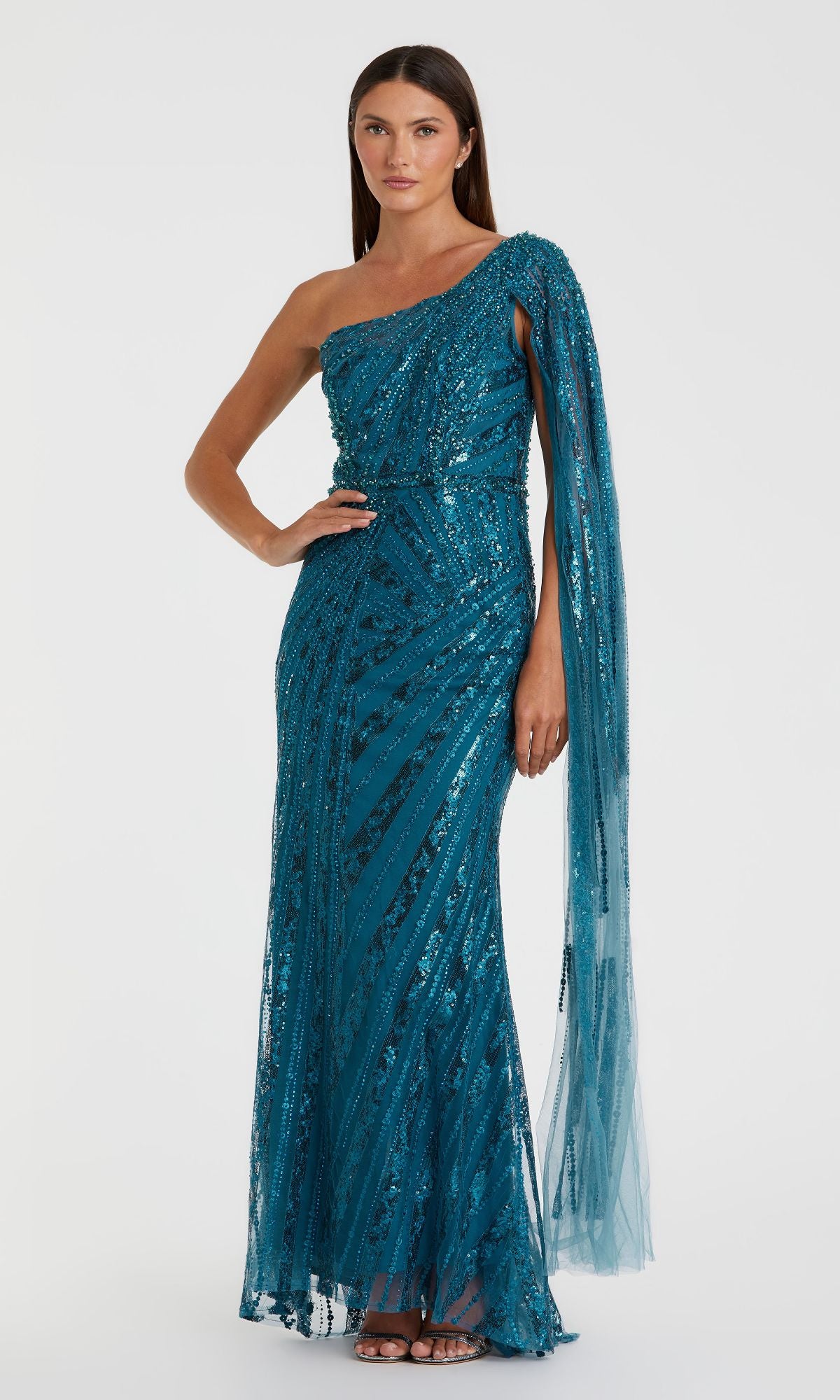 A woman models the Mac Duggal 20528 One-Shoulder Long Prom Gown in teal with sequins and a draped sheer sleeve, standing against a plain white background.