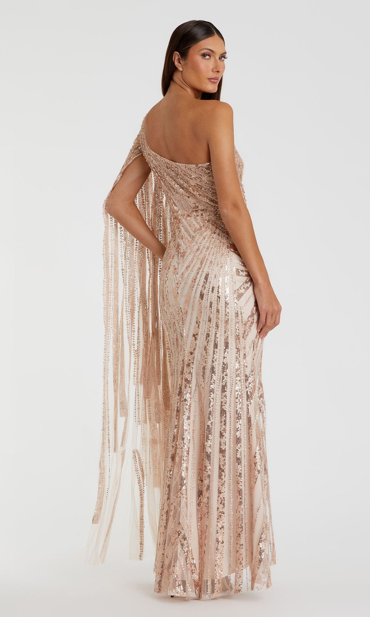 A woman models the Mac Duggal 20528 one-shoulder long prom gown in rose gold sequins with vertical stripes and fringe on one sleeve, standing against a plain white background.