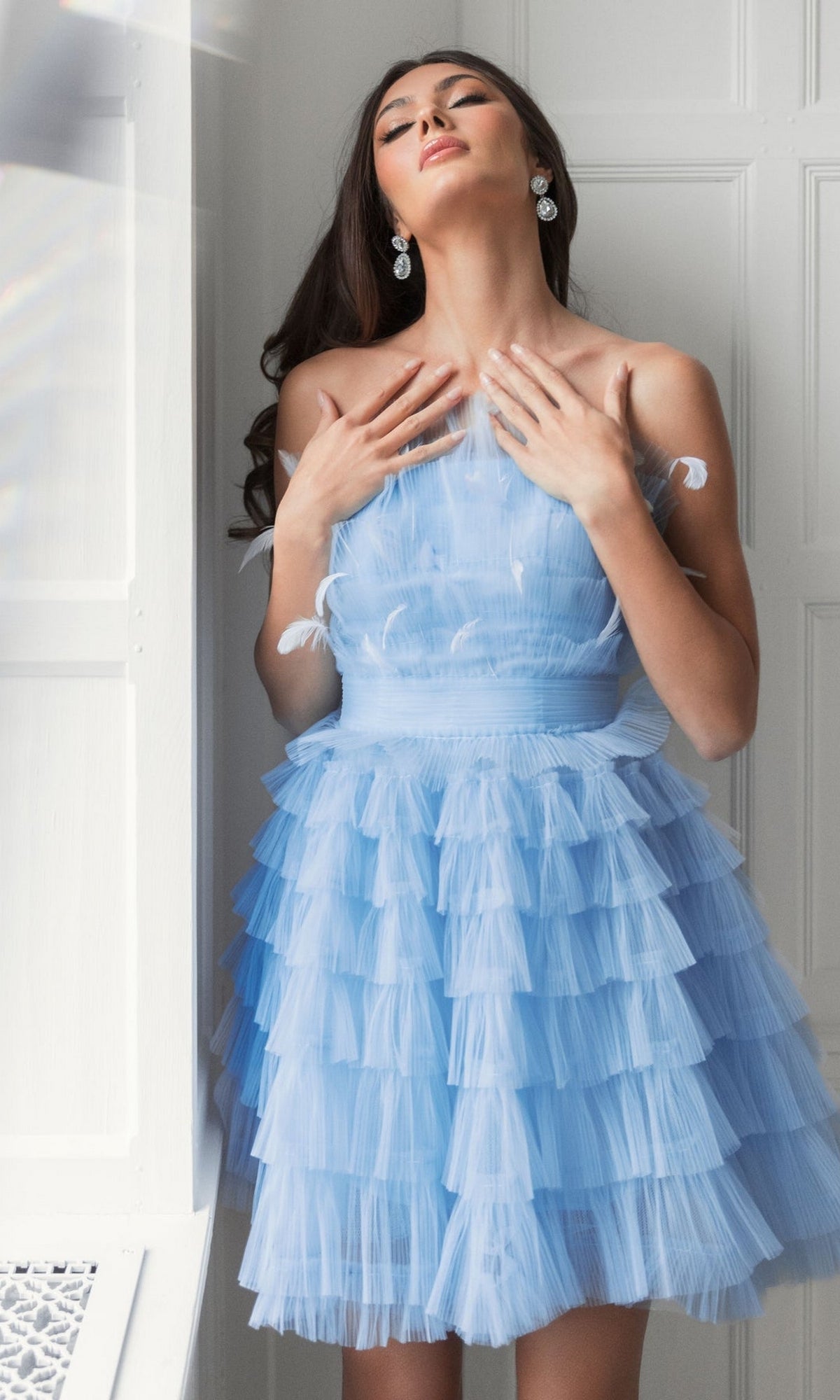 A woman stands by a window in a light blue Mac Duggal 20529 strapless ruffle party dress with feather details, her hands on her chest and eyes closed.