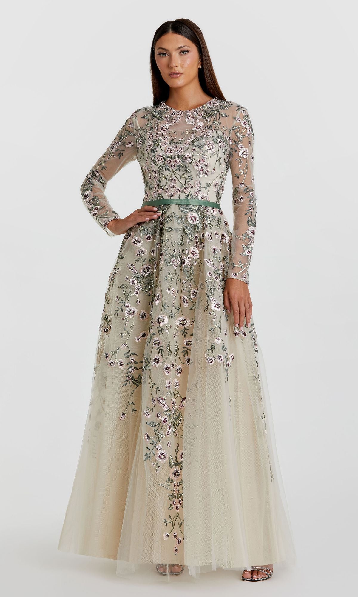 A woman wears the Sage Long Sleeve Formal Gown (Mac Duggal 20580), a sage green floor-length dress featuring sheer sleeves, embroidered floral overlay, and a matching waistband for an elegant look.