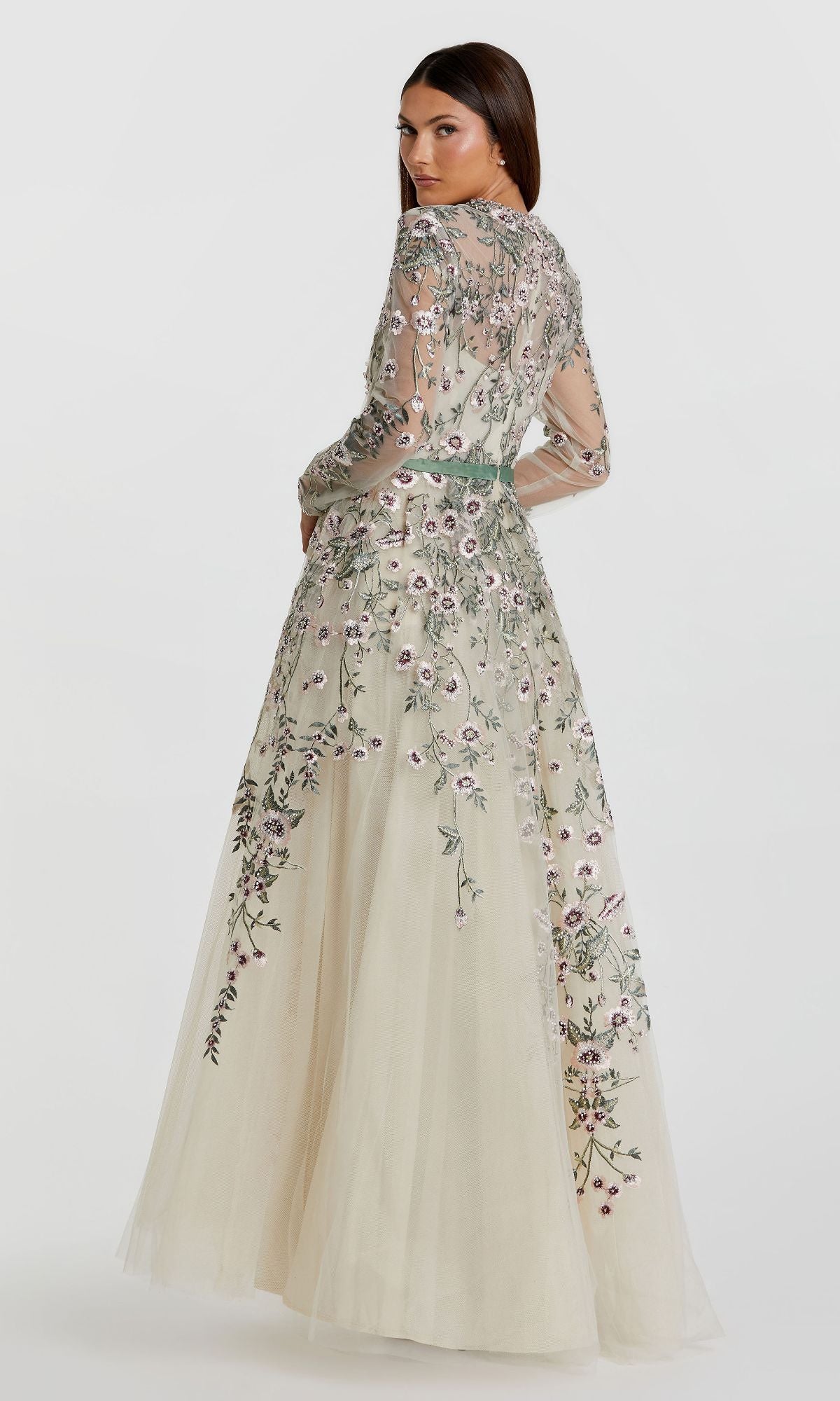 A woman in the Sage Long Sleeve Formal Gown by Mac Duggal (20580), a sheer, cream dress with embroidered flowers, stands facing away and glances over her shoulder against a plain white background.