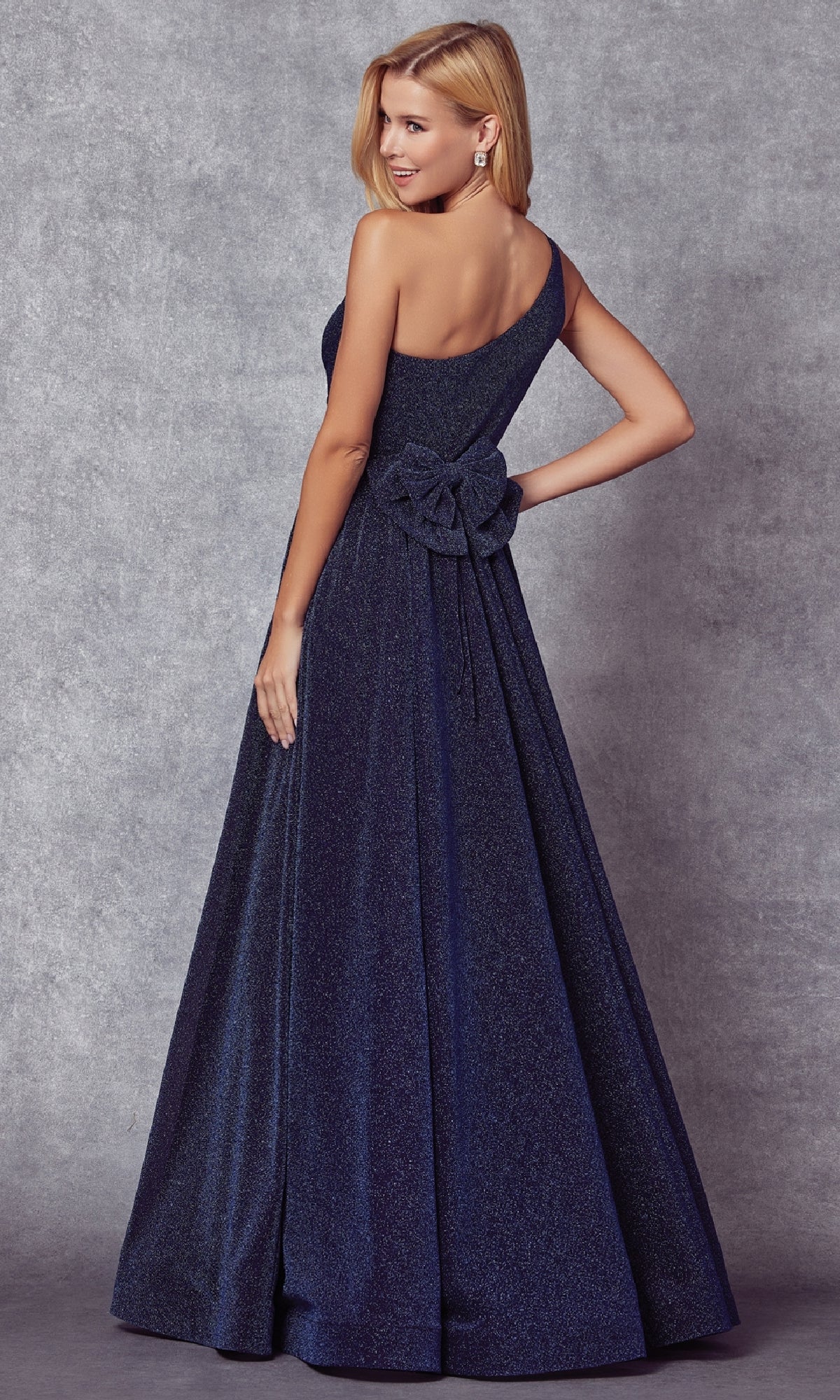 A woman wears the Juliet 205 long navy blue prom dress, featuring a floor-length one-shoulder design and a large bow at the waist, posed against a gray textured backdrop.