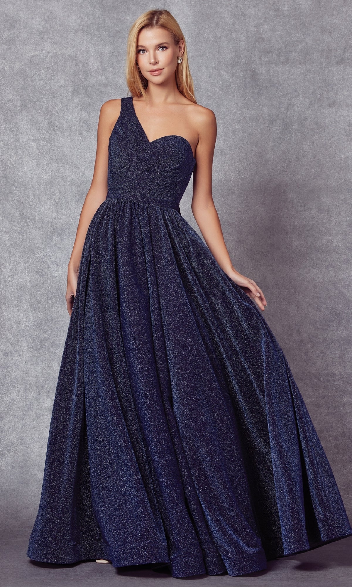 A woman in the Juliet 205 long prom dress, featuring a dark blue one-shoulder design and elegant floor-length silhouette, stands gracefully against a gray textured background.