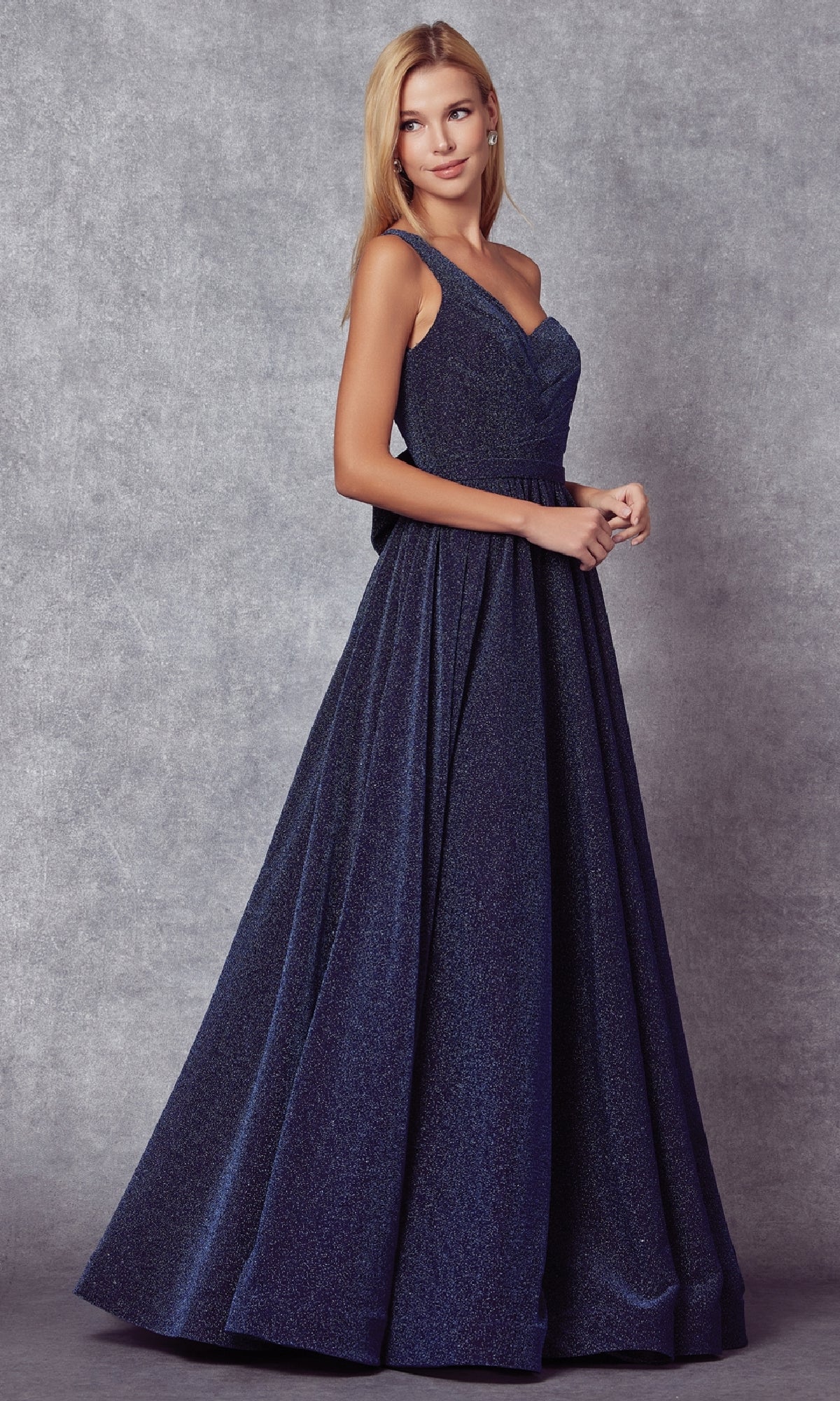 A woman stands before a gray textured backdrop, wearing the Juliet 205 Long Prom Dress—a floor-length, navy blue, one-shoulder gown with an elegant flared skirt.