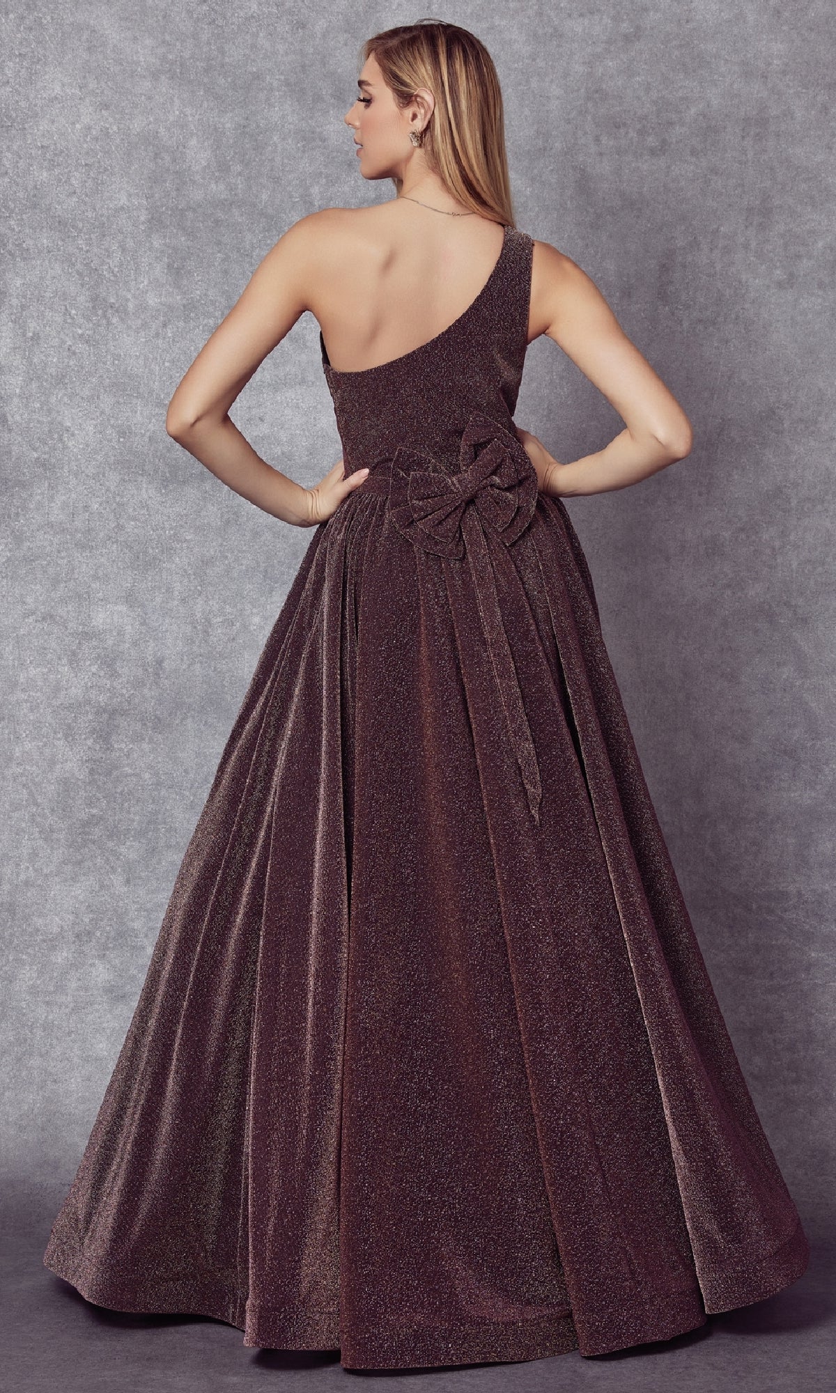 A woman faces away from the camera in the Juliet 205 long prom dress, featuring a shimmery brown, floor-length one-shoulder design with a large waist bow, set against a gray textured background.