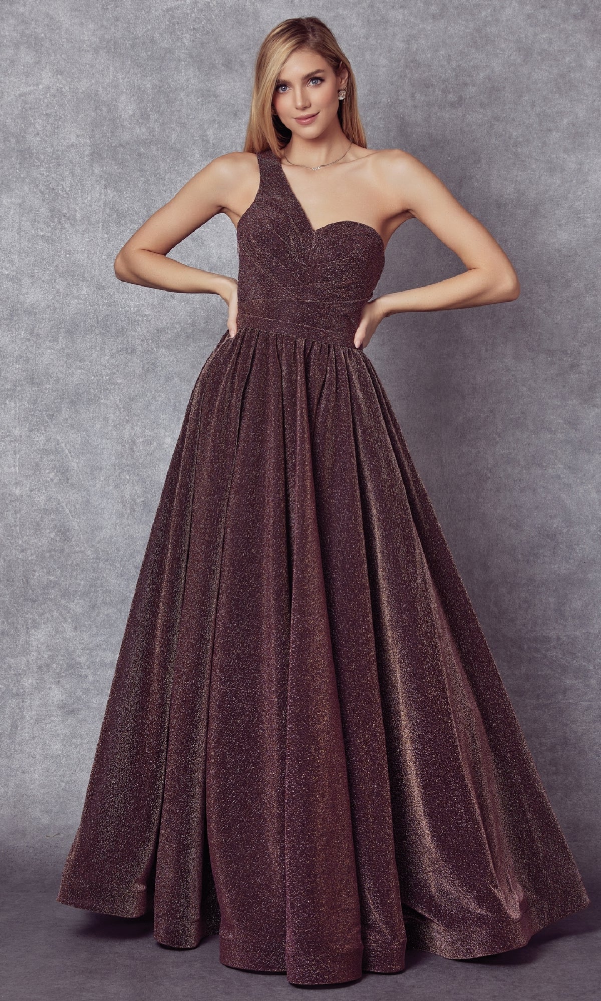 A woman stands against a gray backdrop, wearing the Juliet 205 long prom dress featuring a one-shoulder neckline and a full A-line skirt with glittering fabric.