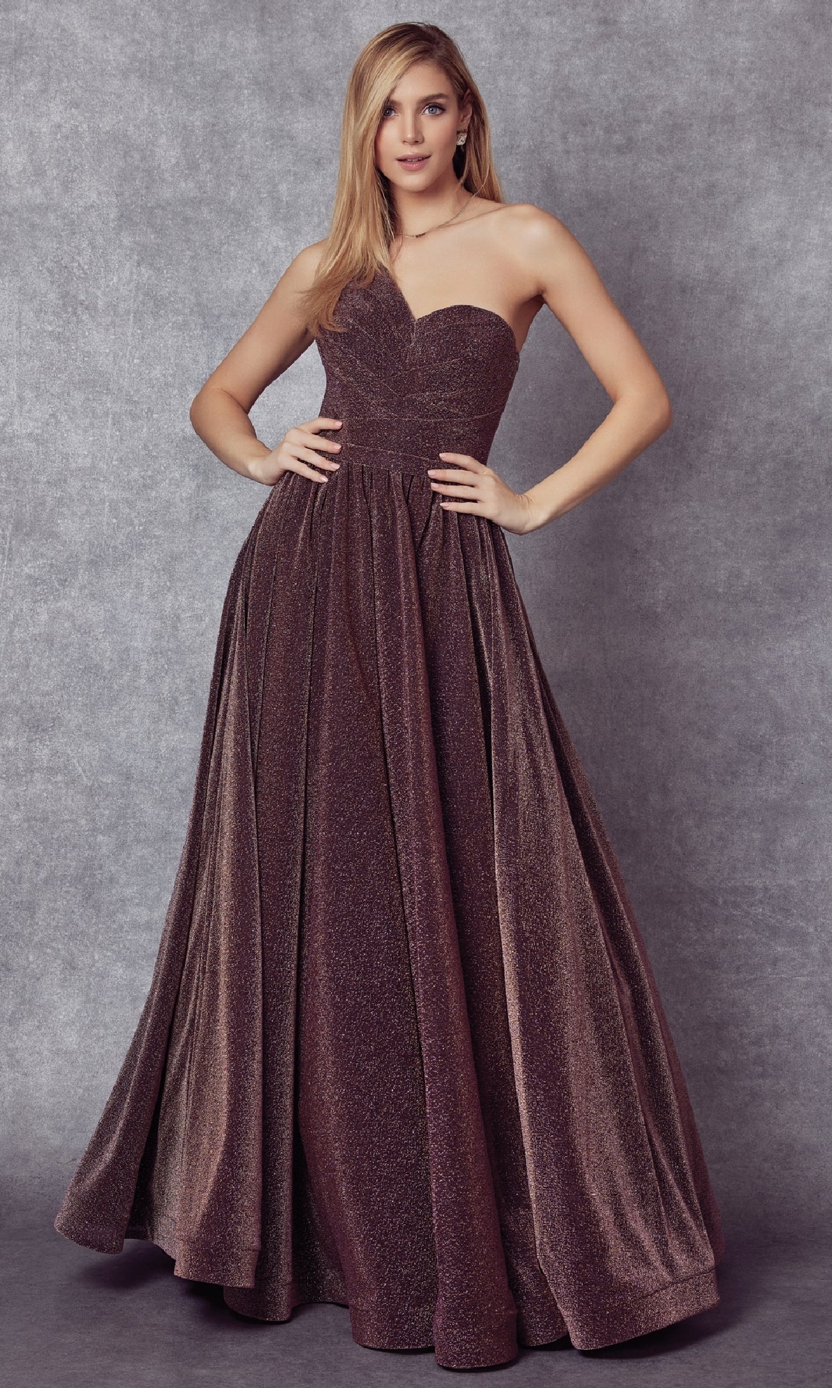 A woman poses against a gray backdrop, wearing the Juliet 205—a floor-length prom dress featuring a strapless fitted bodice and a full, glittering skirt.