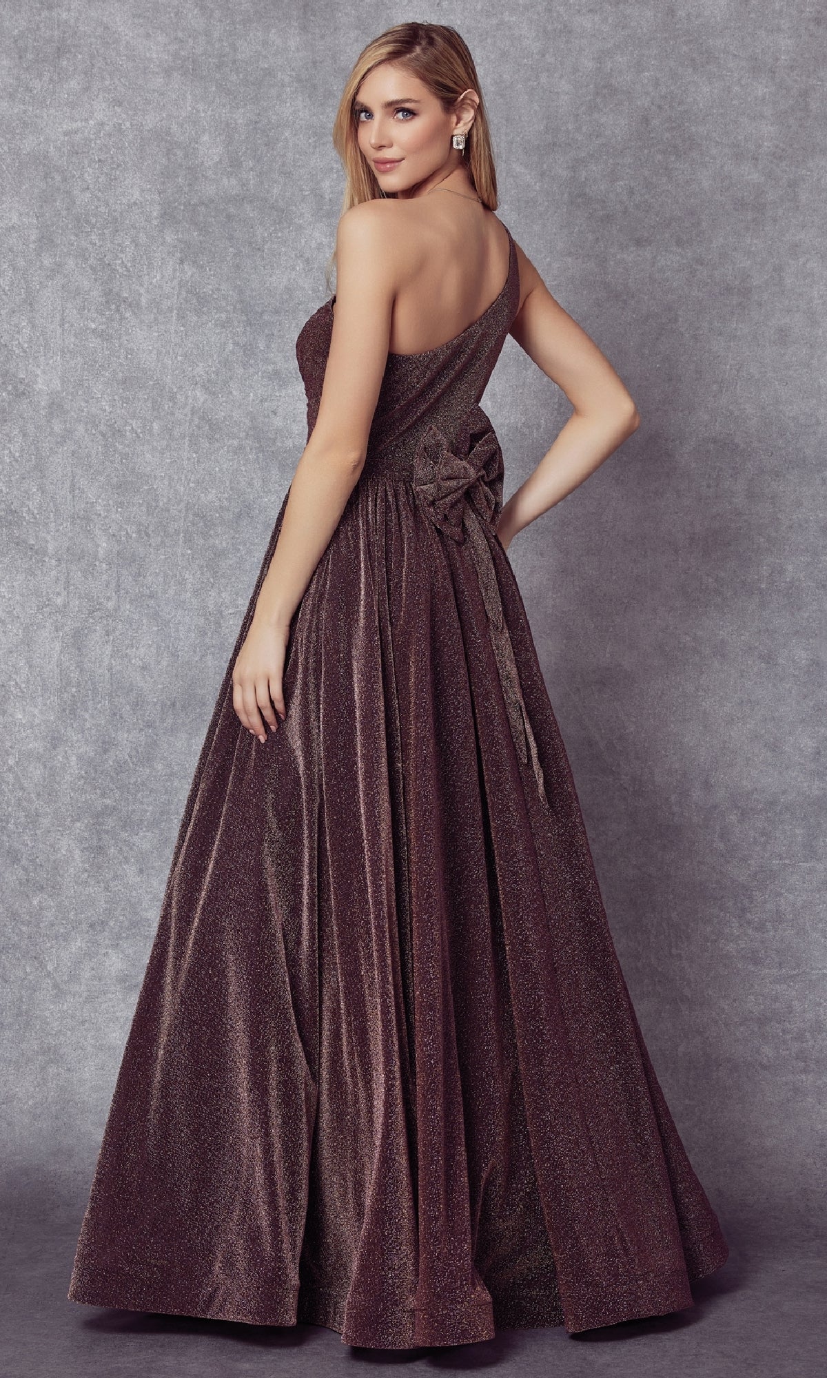 A woman wears the Juliet 205 long prom dress, a floor-length shimmery brown gown with an off-shoulder design and large back bow, standing against a gray textured background.