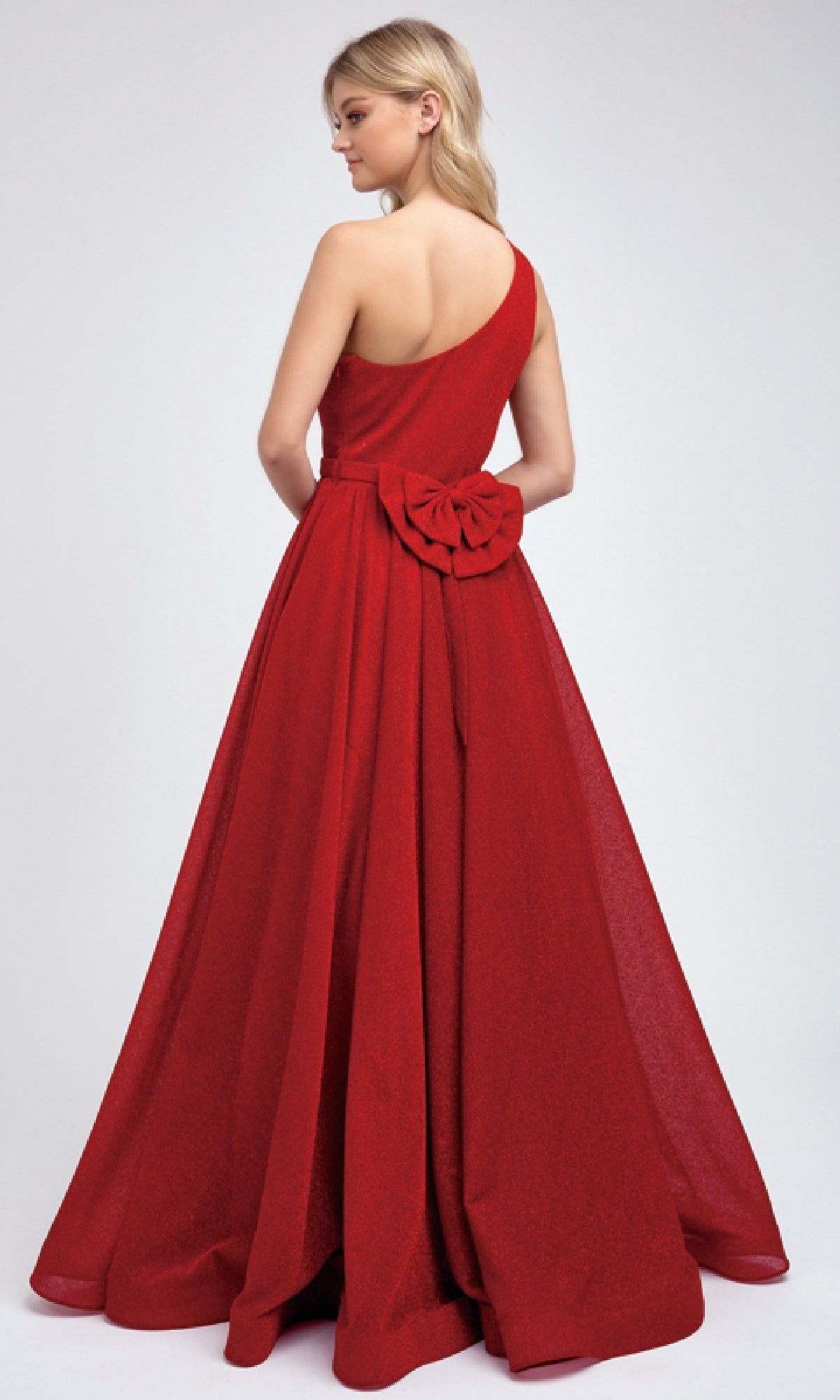 A woman stands facing away in the Juliet 205 long prom dress—red, floor-length, one-shoulder style with a large flower detail at the waist.