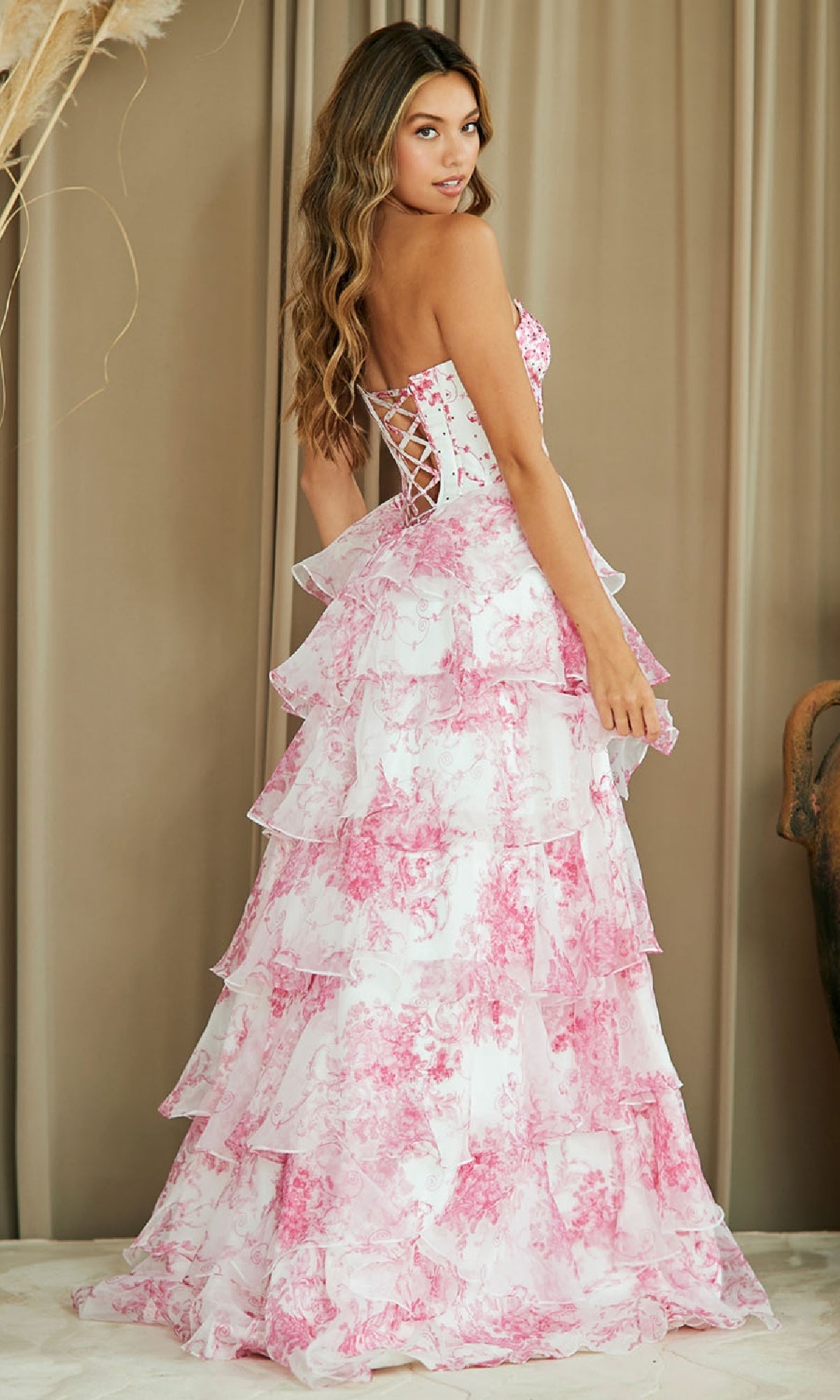 Woman wearing the Strapless Print Prom Gown: Amelia Couture 2060, a strapless long floral dress with a corset-style bodice and pink and white tiered layers, stands in front of beige curtains.
