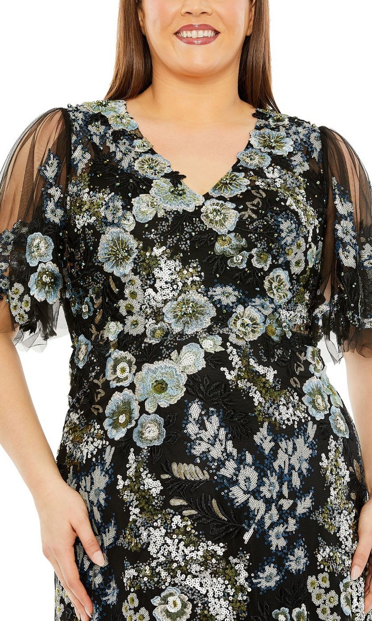 A woman wears the Long Plus-Size Prom Dress 20630 by Mac Duggal, featuring black fabric, sheer short sleeves, and an intricate floral sequin pattern in blue, white, and green.