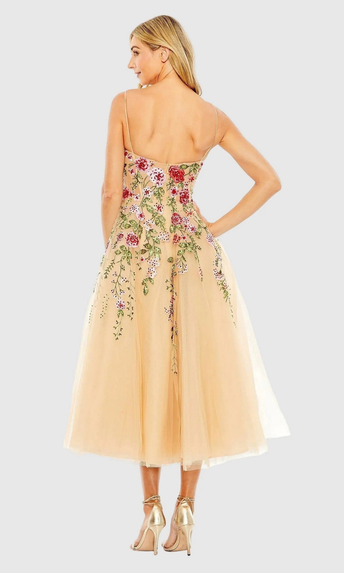 A woman faces away, wearing the Mac Duggal 20651 Floral Embroidered A-Line Midi Dress with spaghetti straps. The light beige cocktail dress pairs perfectly with her long blonde hair and gold high-heeled sandals.