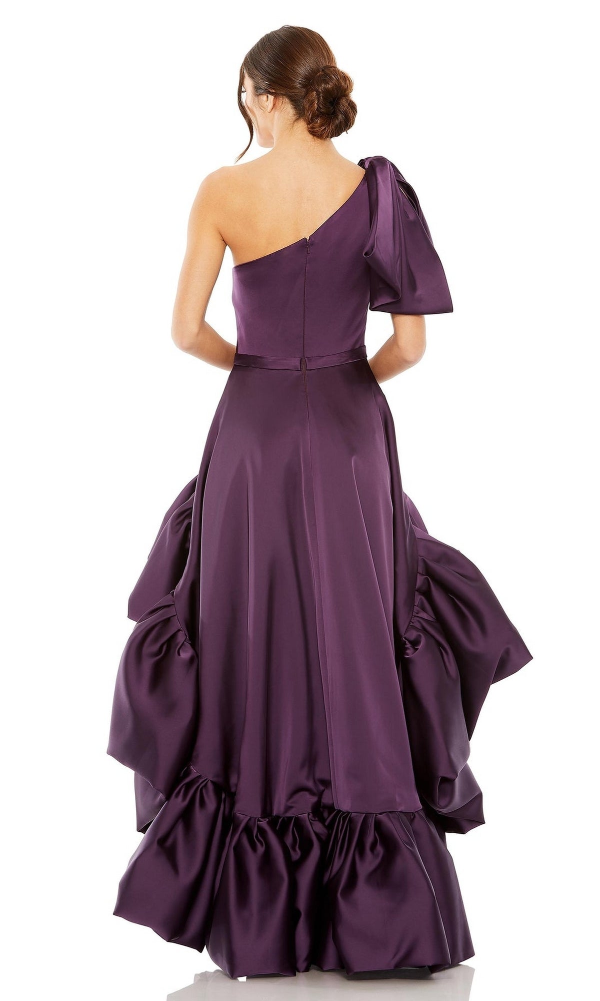A woman stands facing away, wearing the High-Low Party Dress 20702 by Mac Duggal—an aubergine purple, one-shoulder satin gown with ruffled detailing and a shoulder bow, perfect for evening elegance.