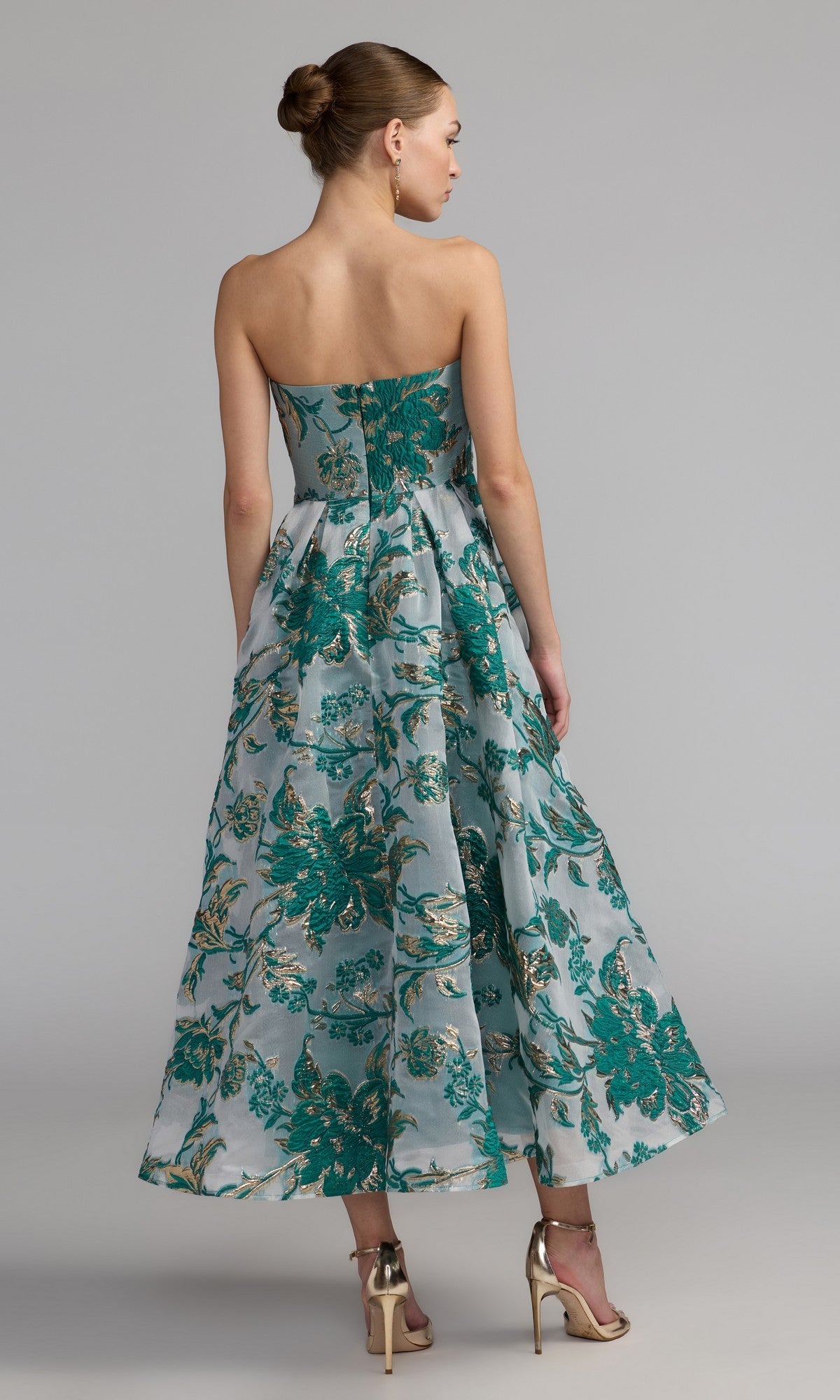A woman in a strapless bronze Mac Duggal 20716 party dress and metallic high-heeled sandals stands with her back to the camera.