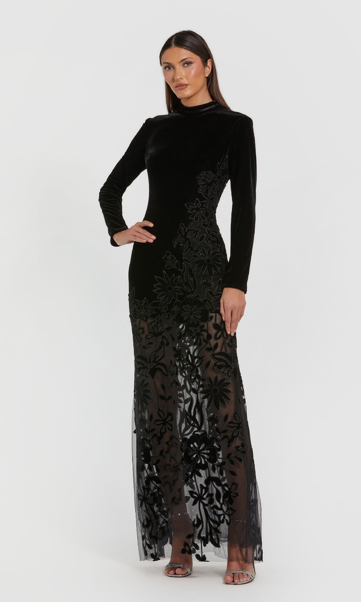 A woman models the Mac Duggal 20746, a long black velvet formal dress featuring floral embroidery and a sheer skirt, posed against a plain white background.