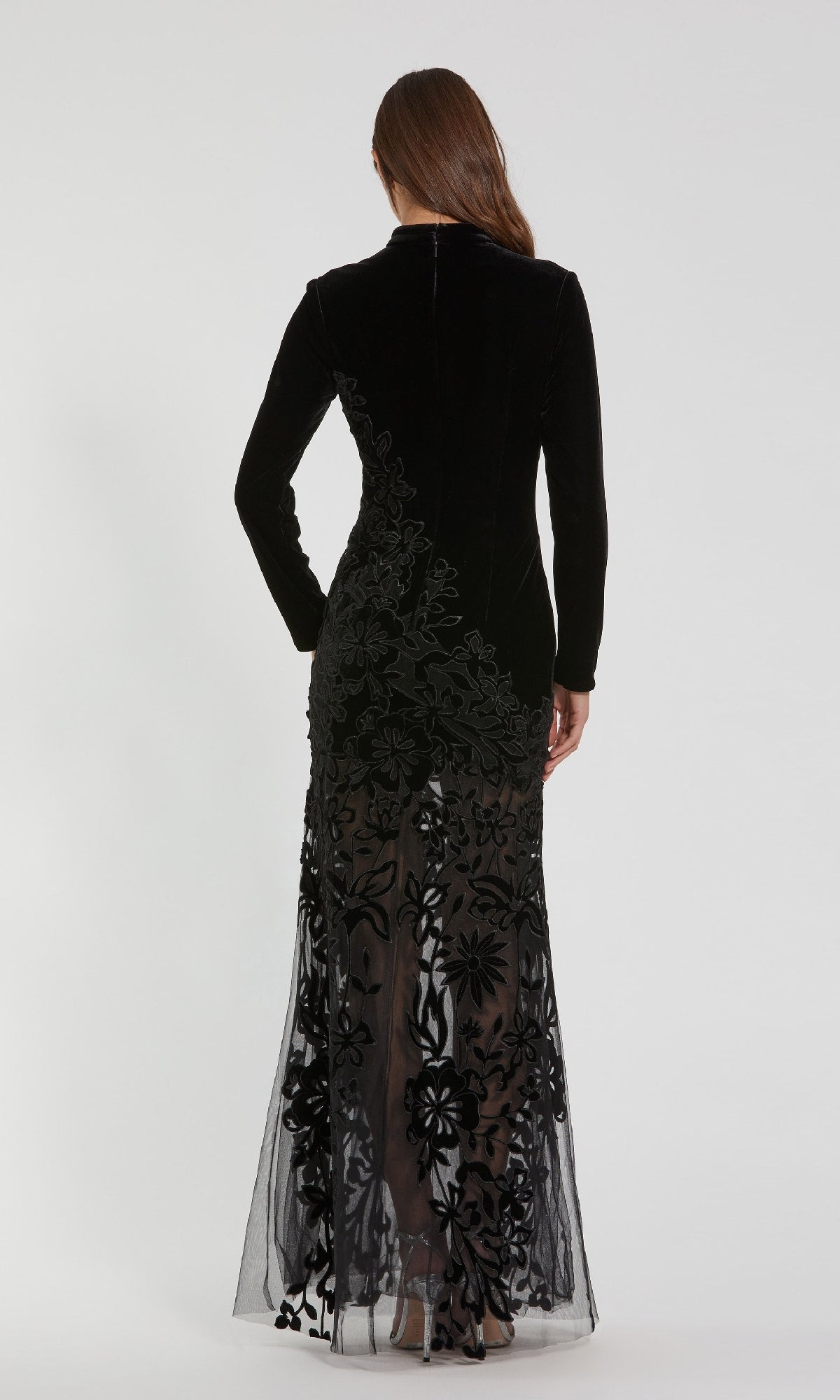 A woman stands facing away, wearing the Mac Duggal 20746—a long black velvet formal dress featuring elegant long sleeves and sheer floral detailing on the lower half.