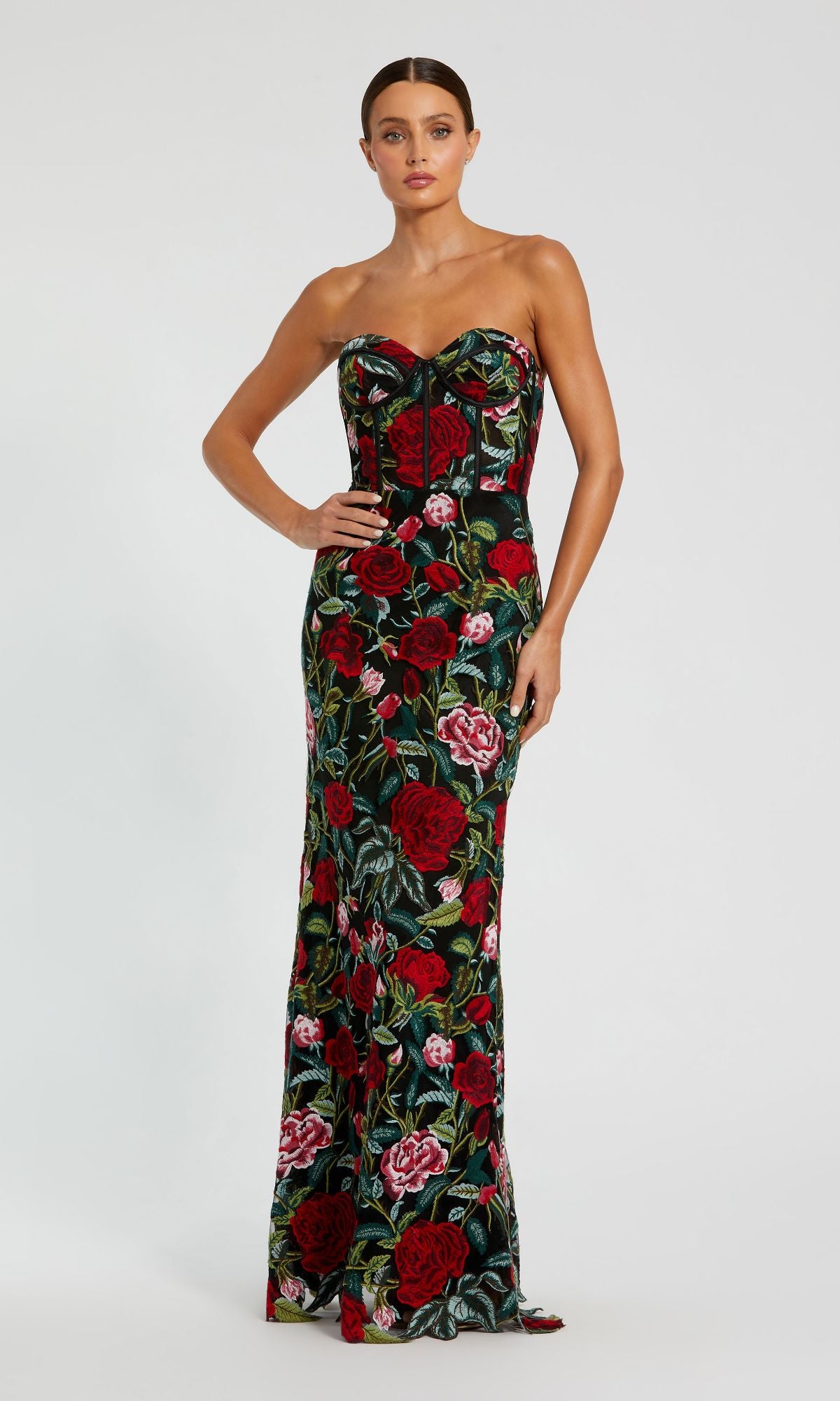 A woman stands gracefully against a plain light backdrop in the Mac Duggal 20757 Long Print Formal Evening Gown, featuring bold red and pink floral prints on a dark background.