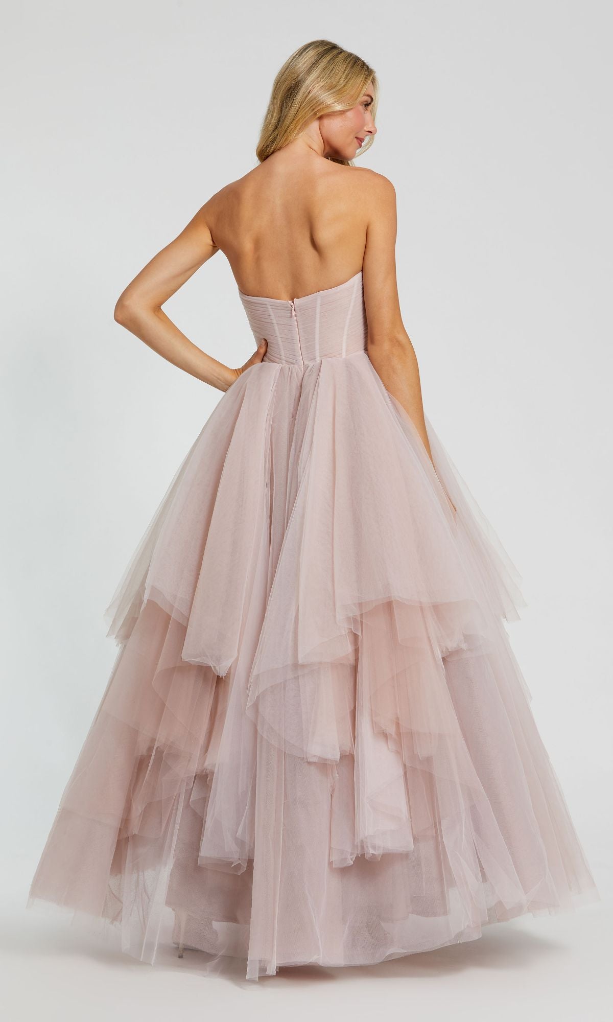 A woman is shown from the back wearing the Long Formal Dress 20772 by Mac Duggal—a strapless blush pink tulle ball gown featuring a ruched bodice and layered skirt—against a plain background.