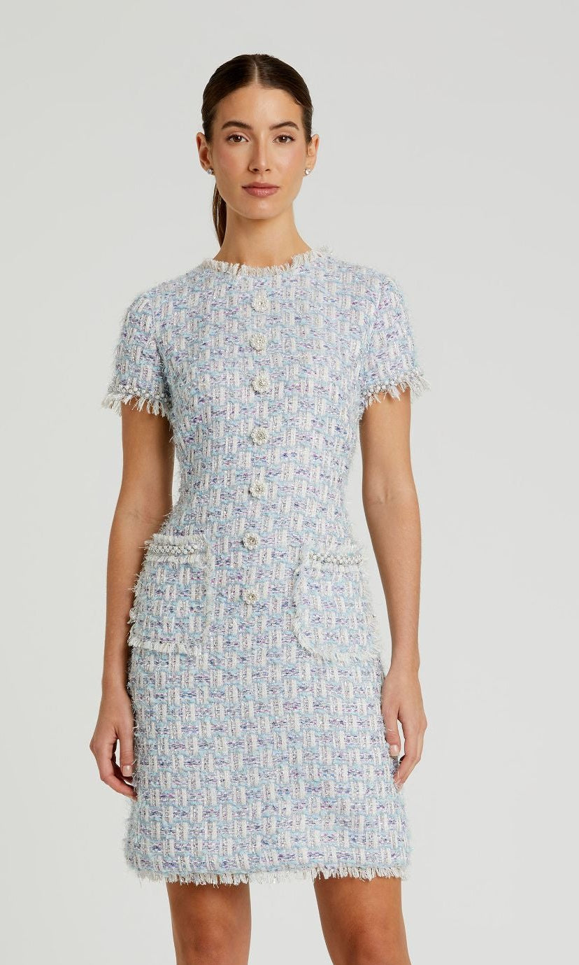 A woman wears the Mac Duggal 20805 Short Sleeve Semi-Formal Dress in light blue boucle with front buttons and two pockets, standing against a plain background.