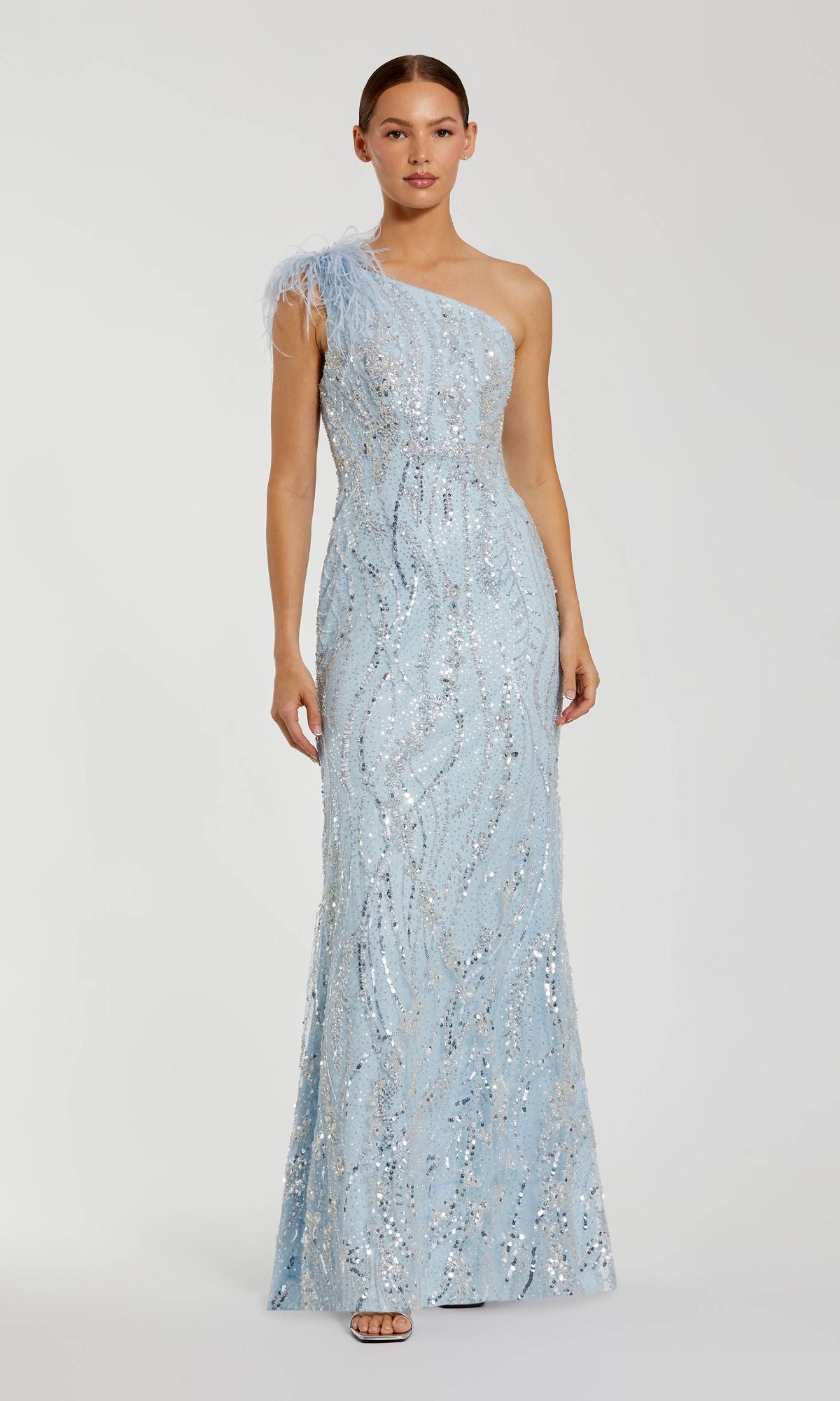 A woman models the Mac Duggal 20887, a long blue glitter formal dress with a one-shoulder design, feather details, and floor-length silhouette, standing against a plain background.